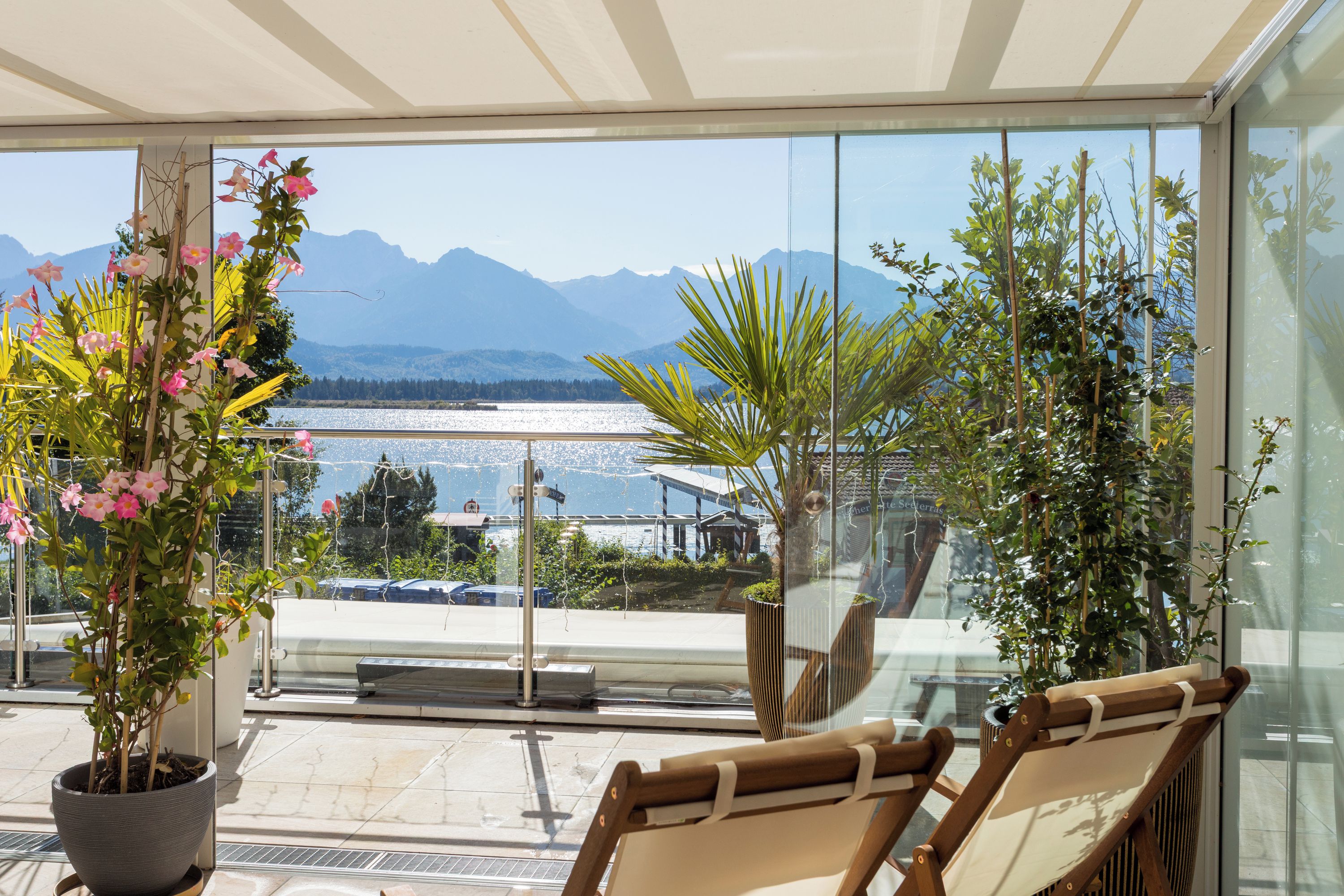 A balcony with a view of mountains and a lake, furnished with chairs and potted plants.