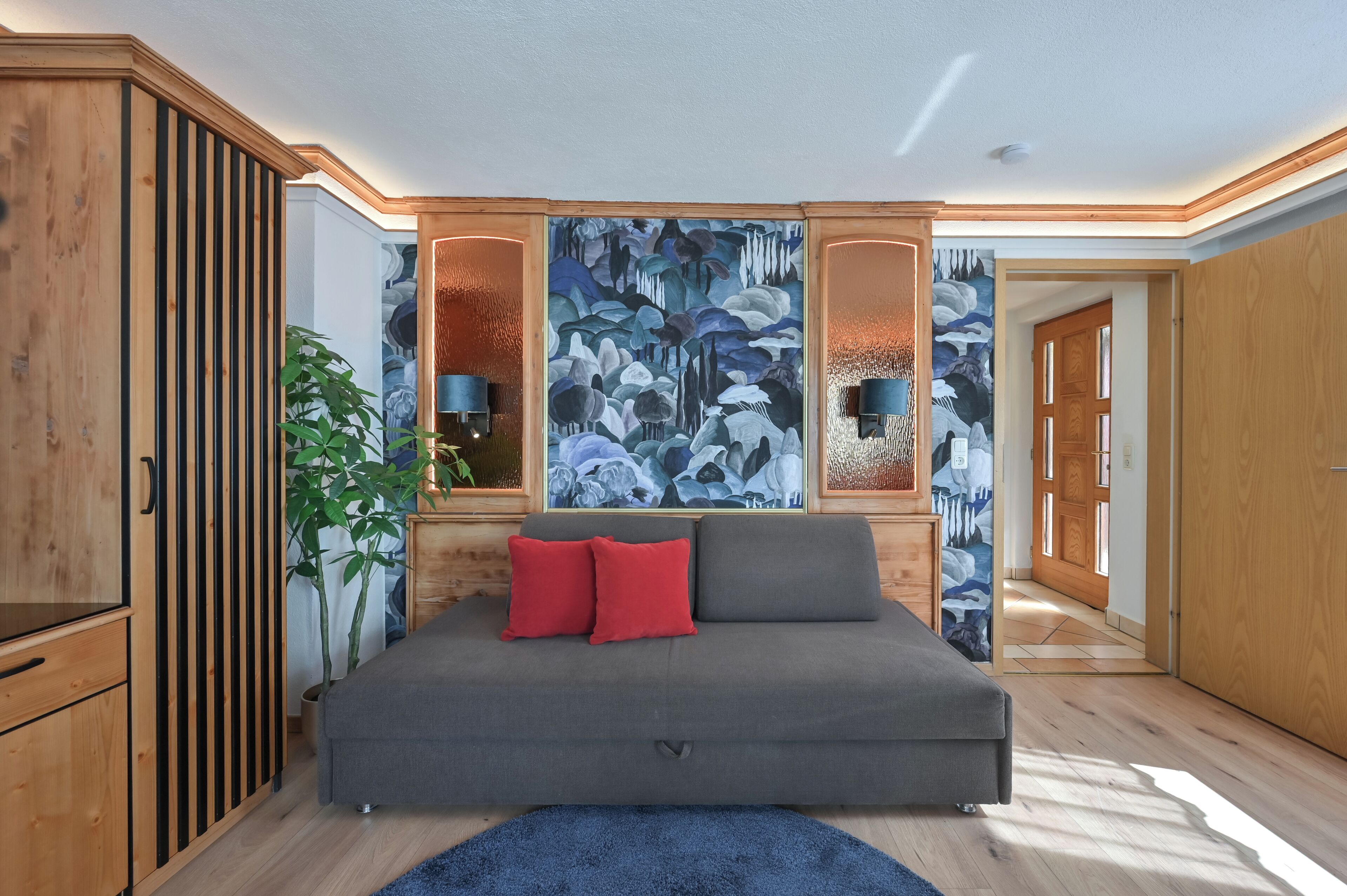 A modern bedroom with a grey sofa bed, a large abstract painting, wooden paneling, and a potted plant.