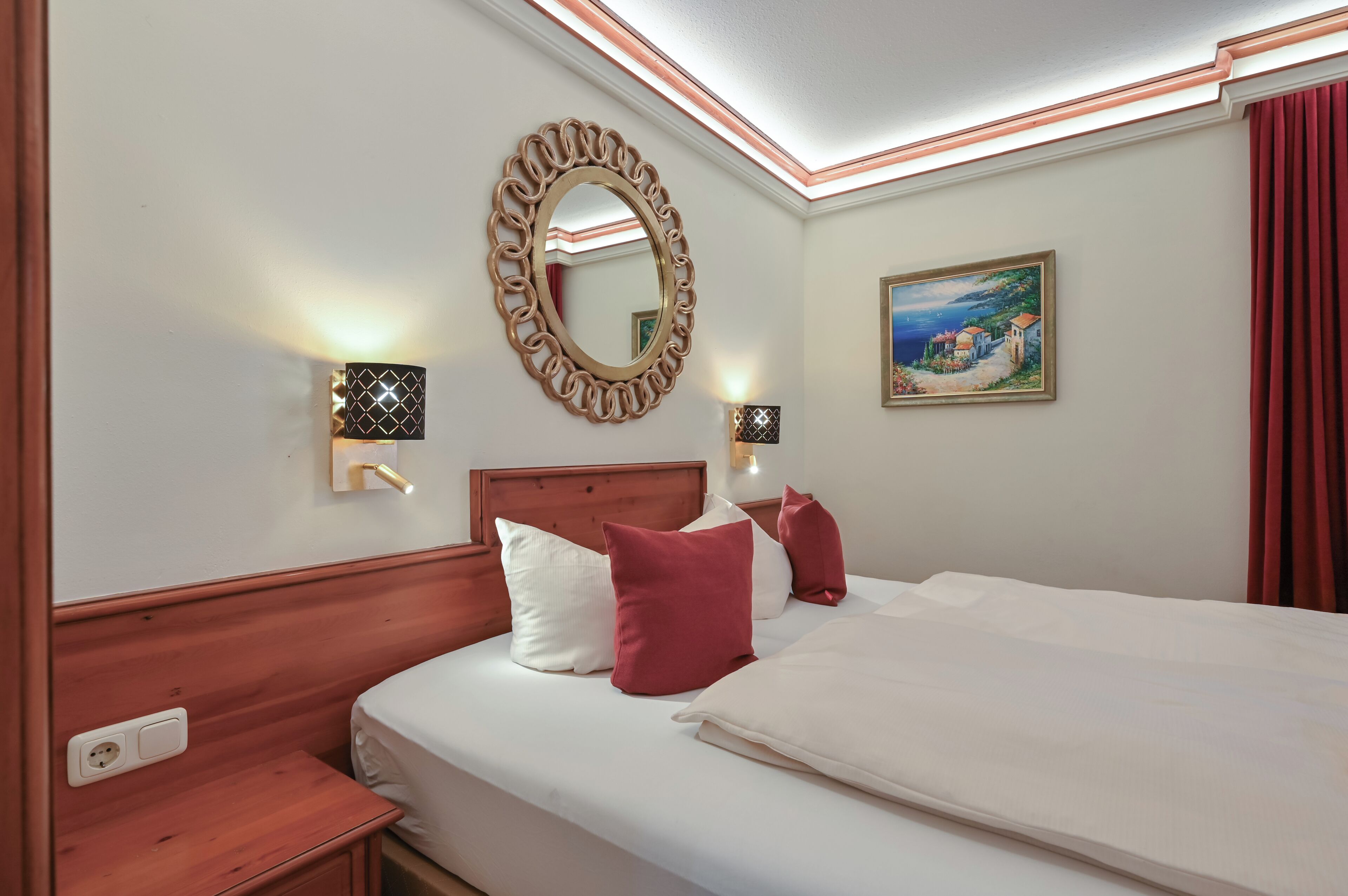 Habitación de hotel con cabecera de madera, cama grande con almohadas blancas y rojas, espejo redondo y dos lámparas de pared.
