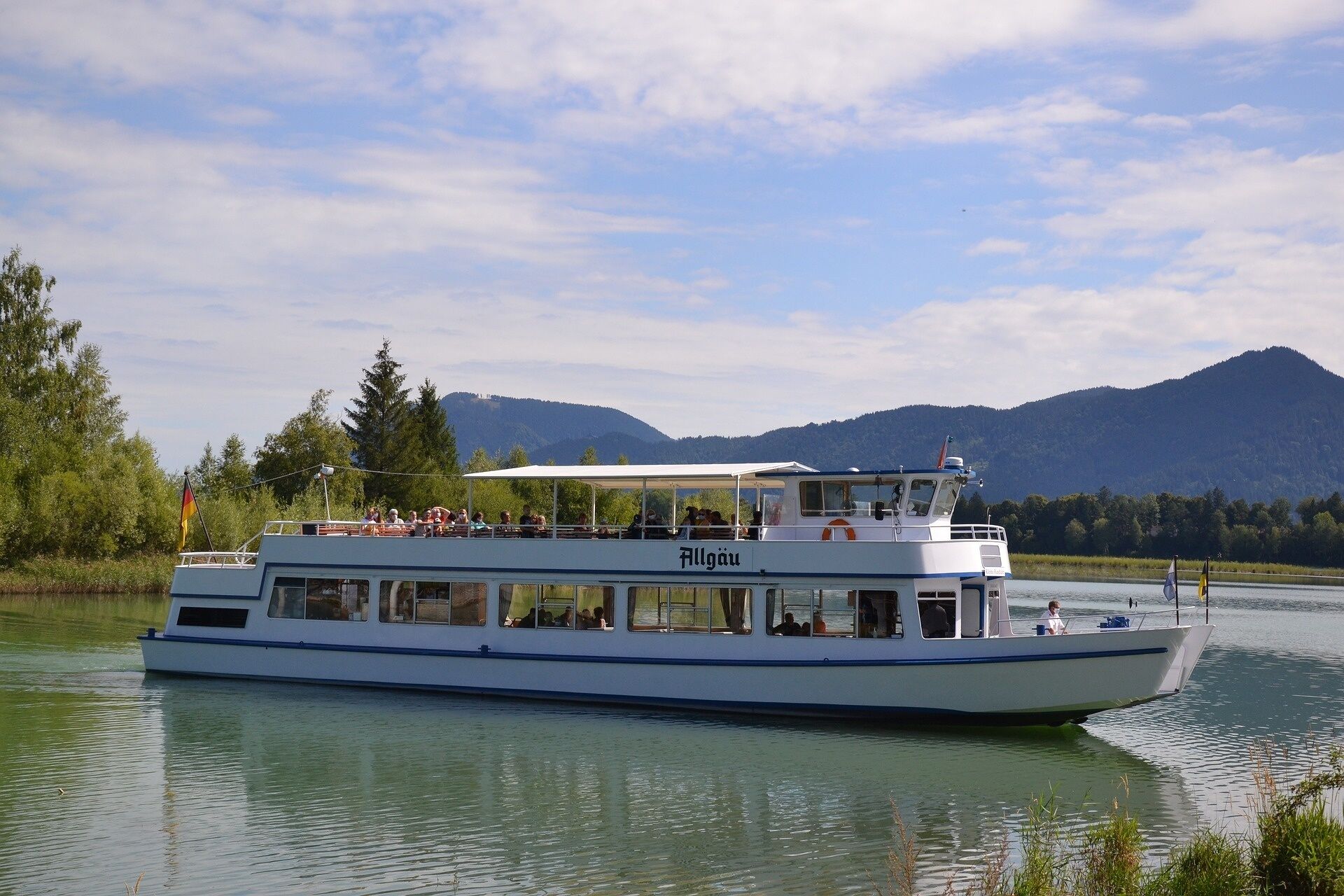 Een boot met de naam 'Allgäu' op de zijkant, vaart over een kalme rivier met een bergachtig landschap op de achtergrond.