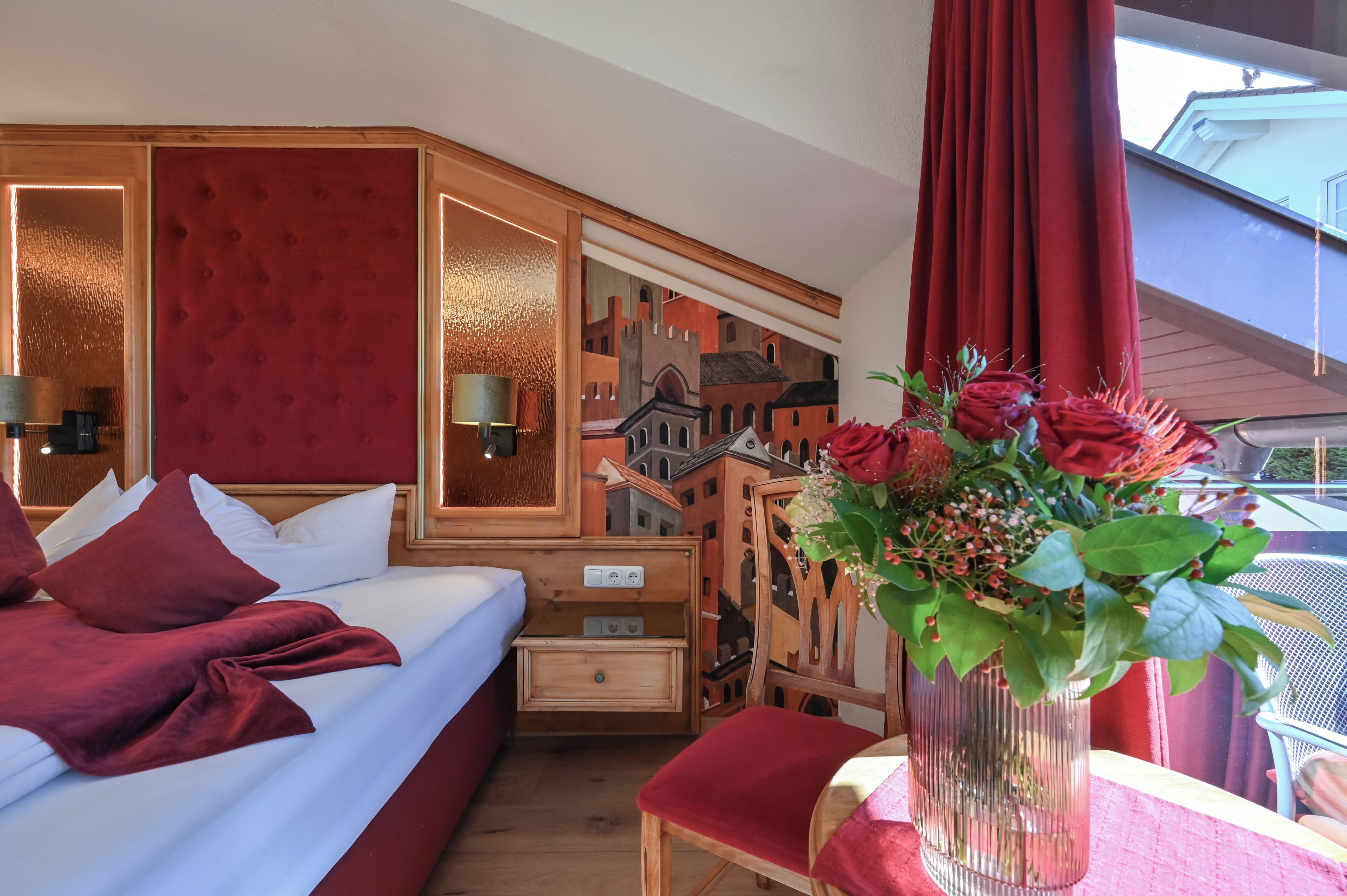 Quarto de hotel com uma cama grande, cabeceira de madeira, mesa de cabeceira, um vaso com flores e vista para um edifício através da janela.