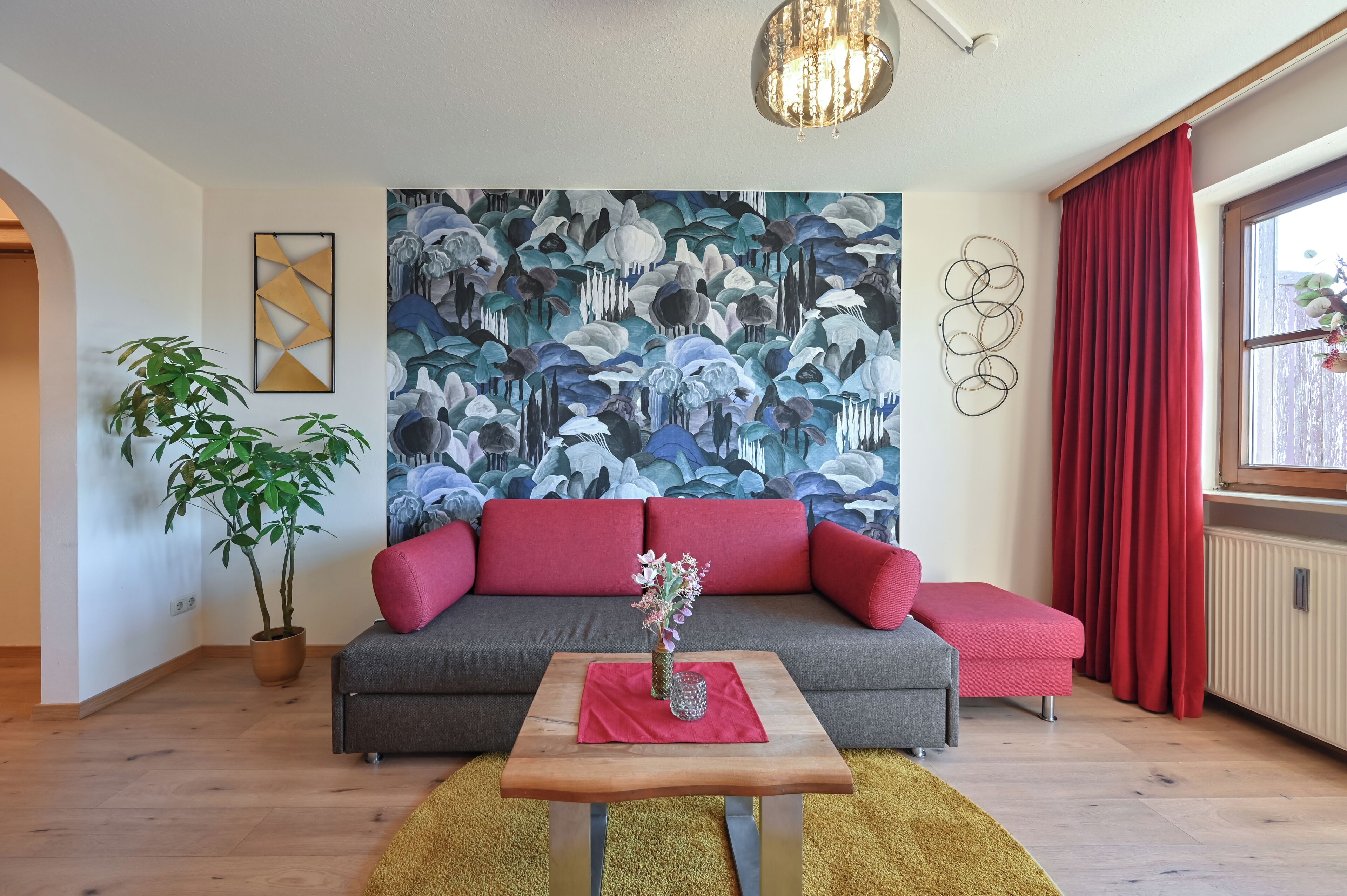 Un salon avec un canapé gris, des coussins rouges, une table basse en bois surmontée d’un vase, une grande fresque murale à motifs floraux, une plante en pot et un rideau rouge.