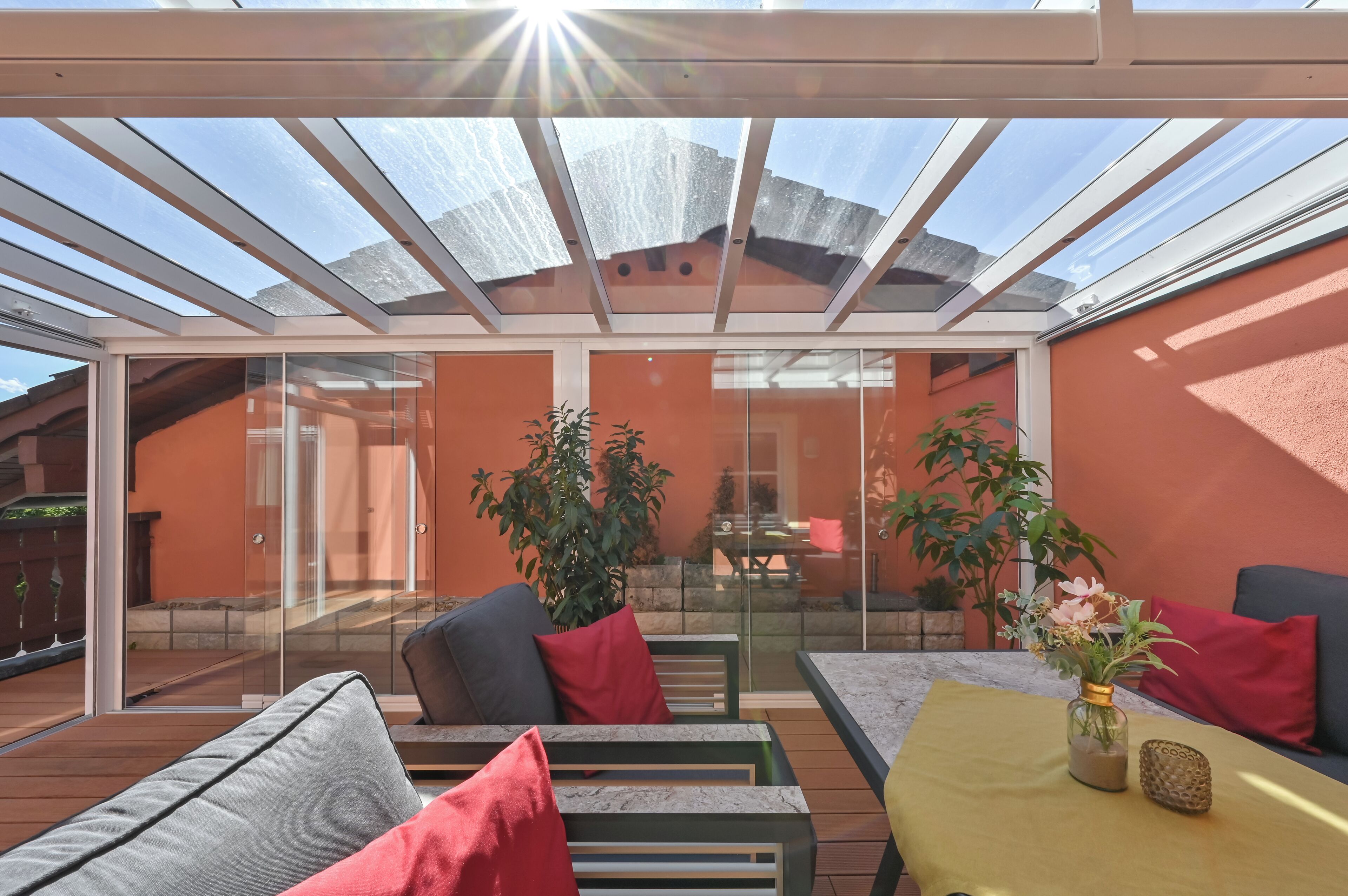 A sunlit patio with a glass enclosure, outdoor seating, and potted plants.