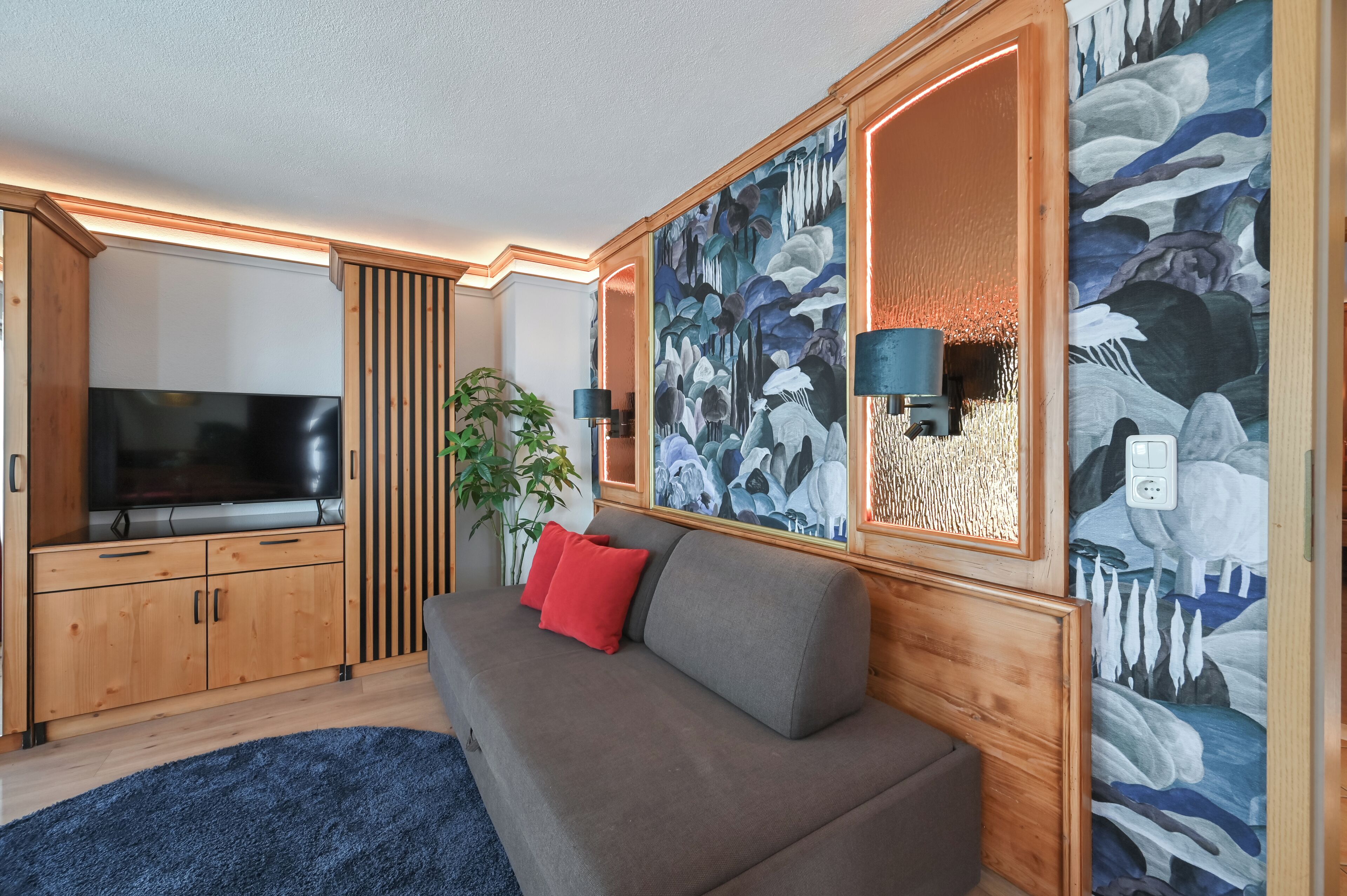 A modern living room with a flat-screen TV, wooden cabinets, a grey sofa with red cushions, and a large abstract painting on the wall.