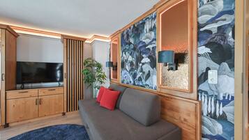 A modern living room with a flat-screen TV, wooden cabinets, a grey sofa with red cushions, and a large abstract painting on the wall.