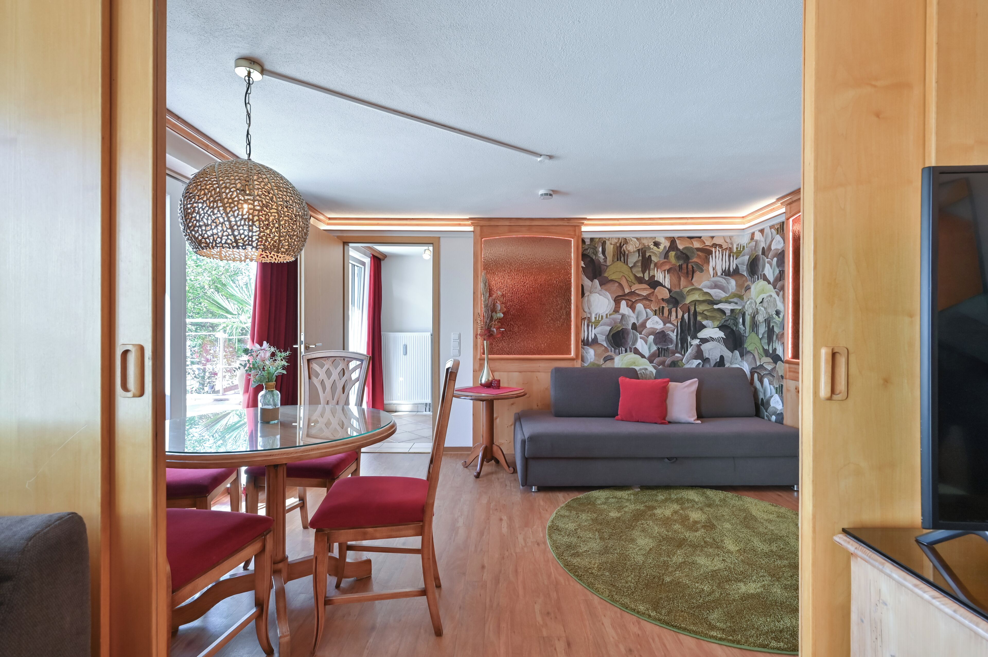 A modern living room with a grey sofa, red cushions, a round glass table with red chairs, a hanging light fixture, and a large abstract wall mural.