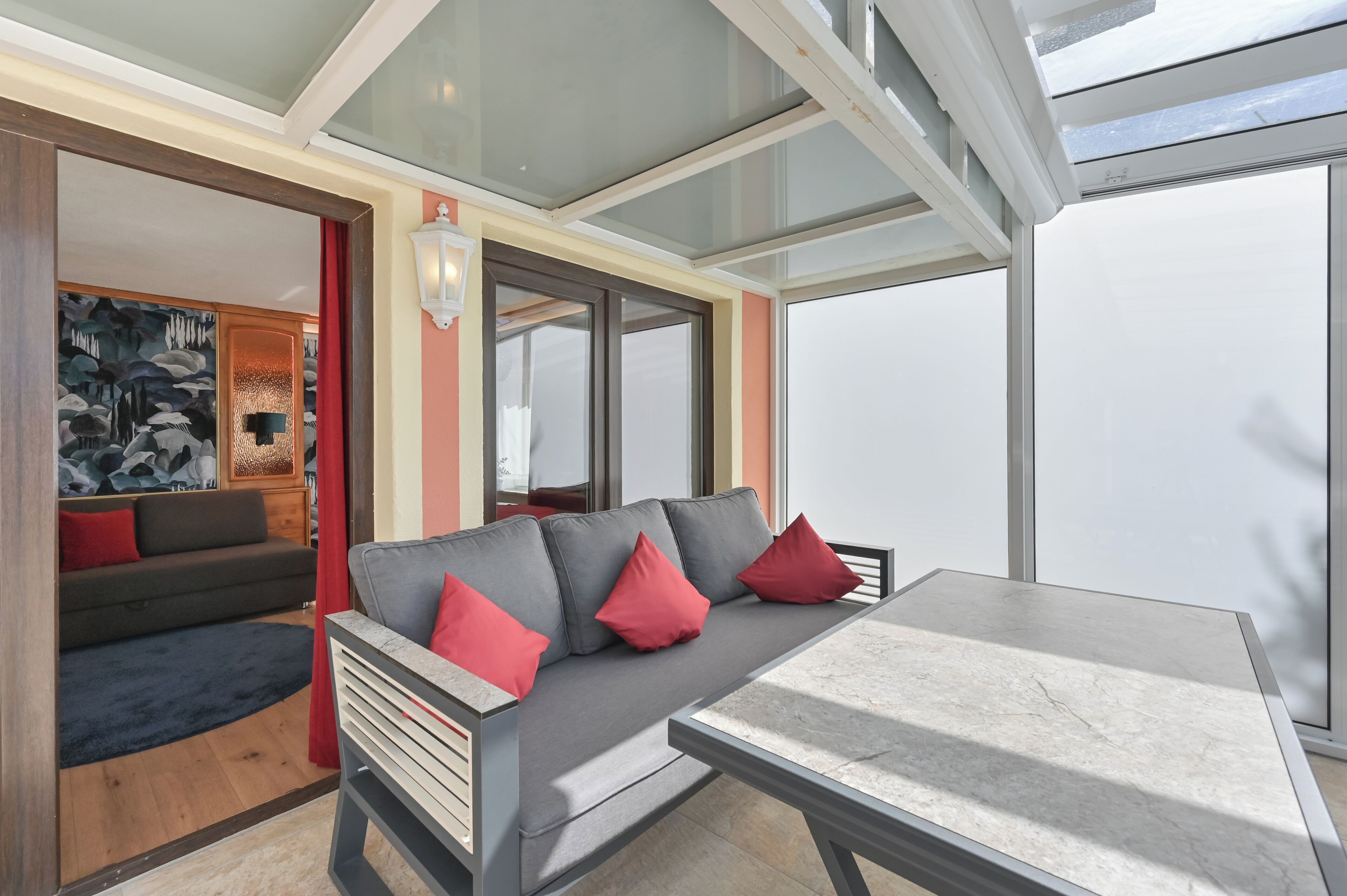 Un interior moderno con un sofá gris, cojines rojos, una mesa de mármol y ventanas amplias.