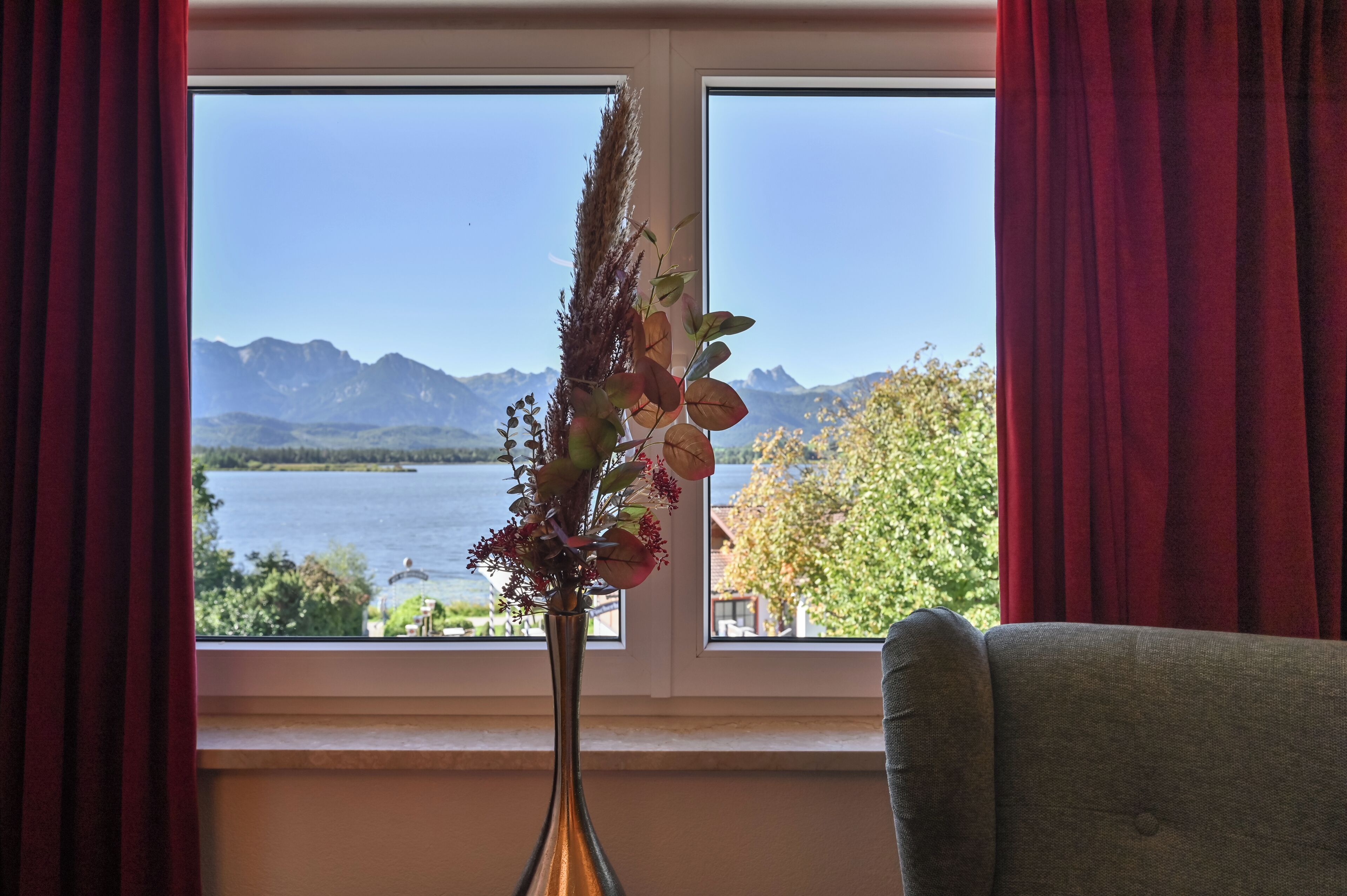 A room with a view of a lake and mountains through a window, a vase with dried plants, and a grey armchair.