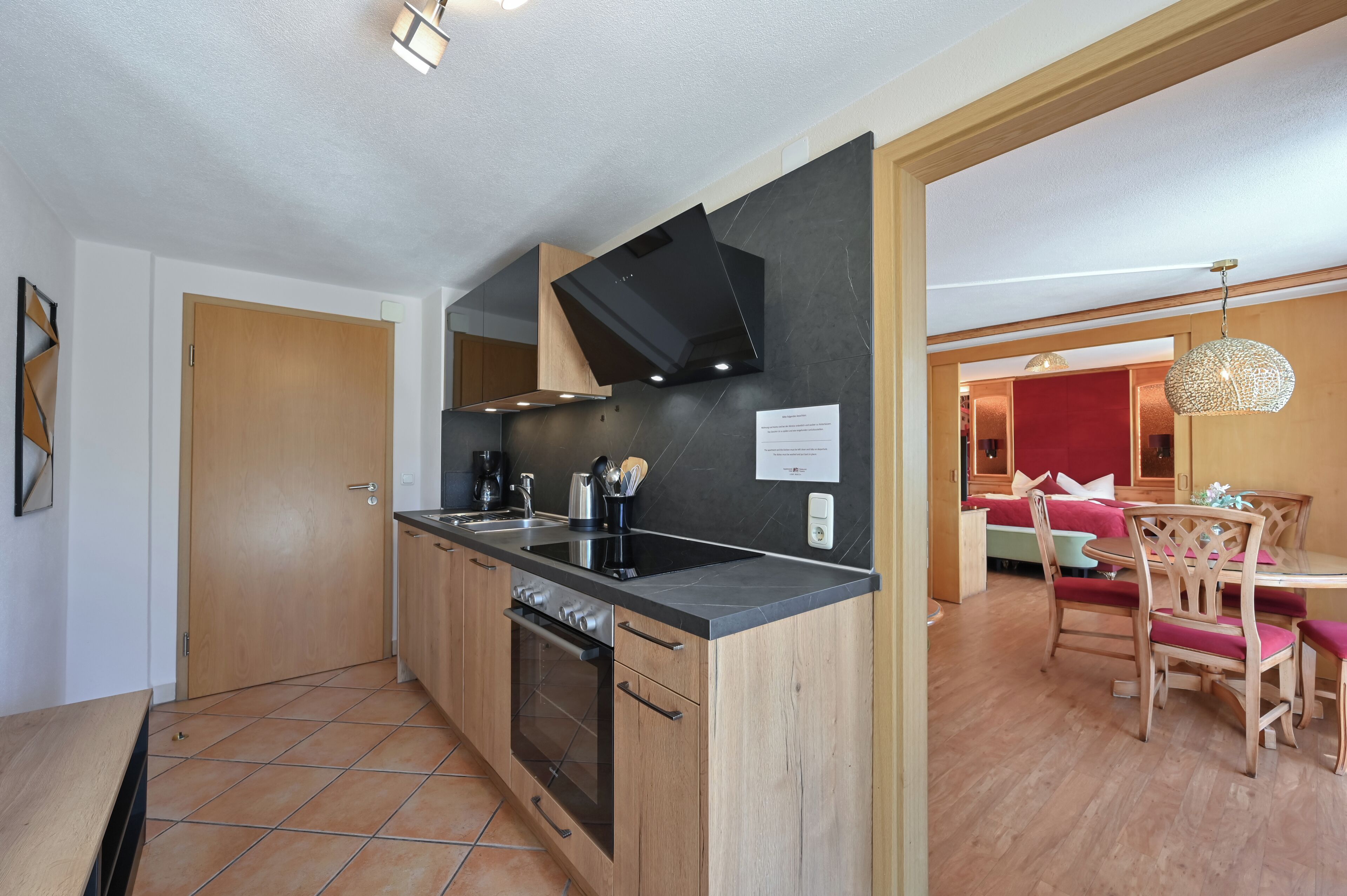 A modern kitchen with a built-in oven, a black range hood, and a dining area with wooden chairs and a red table.