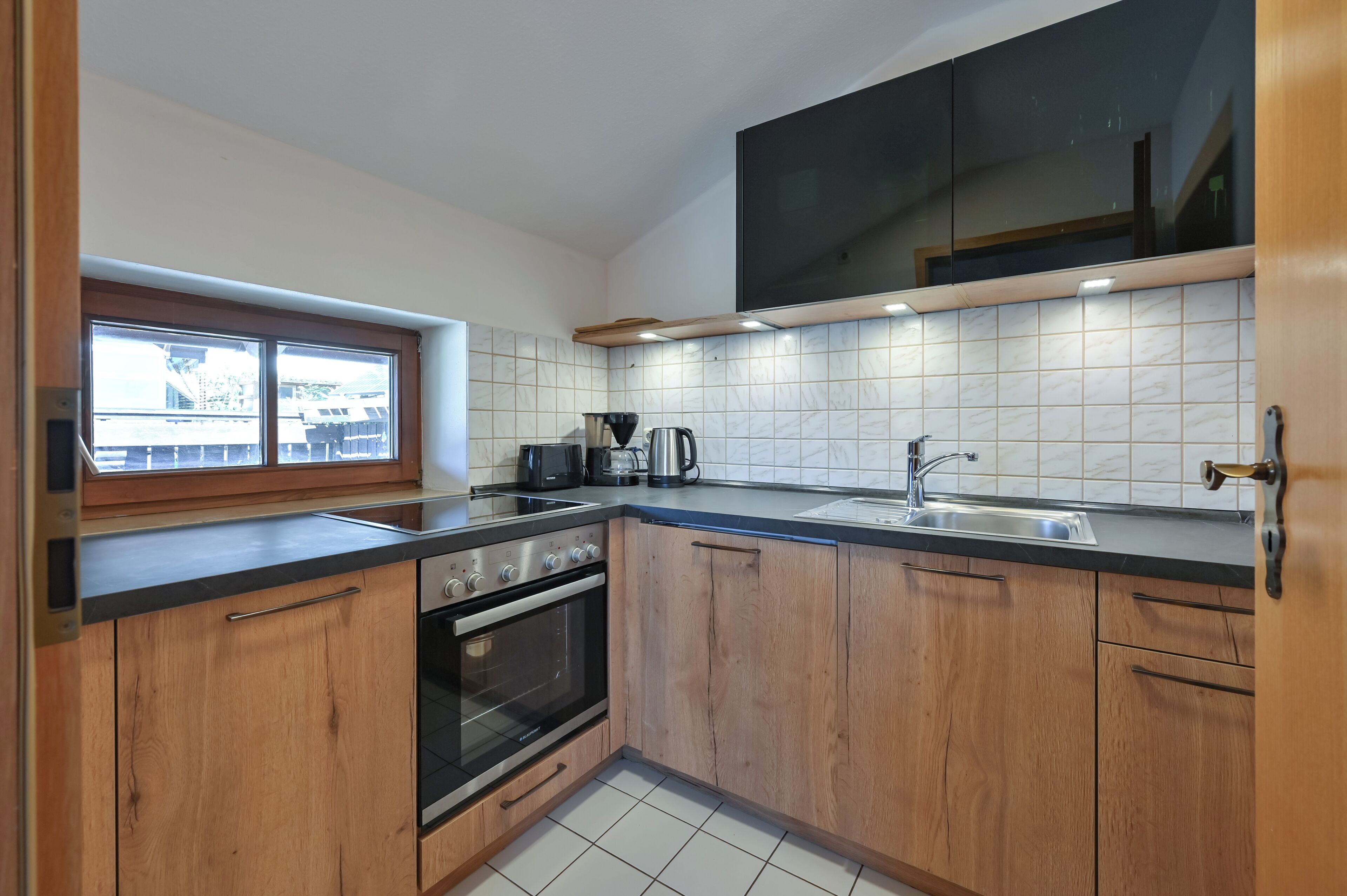 Een keuken met houten kastjes, een inbouwoven, een spoelbak en een raam met uitzicht op gebouwen.