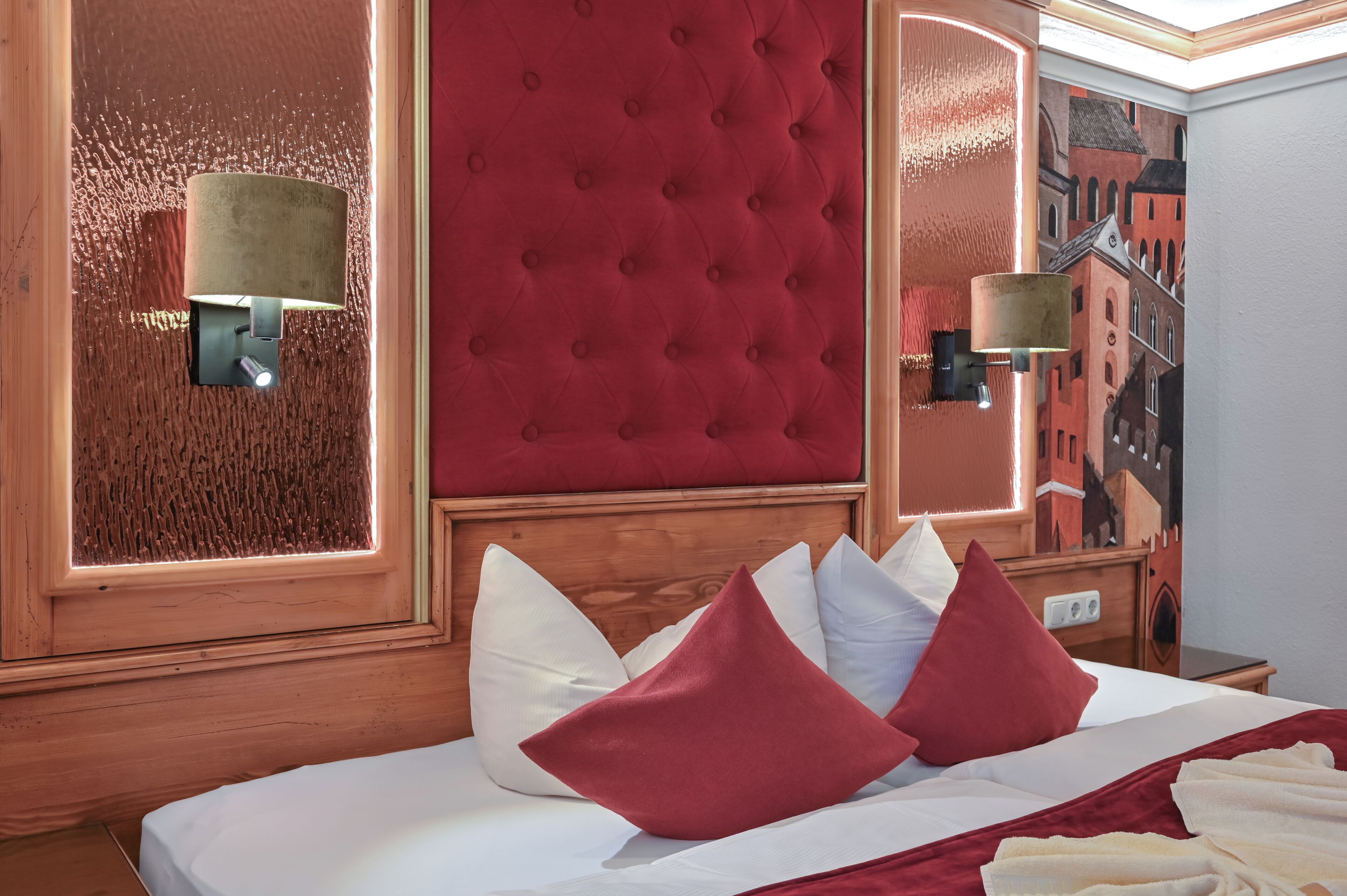 Ein Hotelzimmer mit einer gesteppten Kopfteil, einem Bett mit weiß-roten Kissen, einem Holfnachtkästchen und einer Wandlampe.