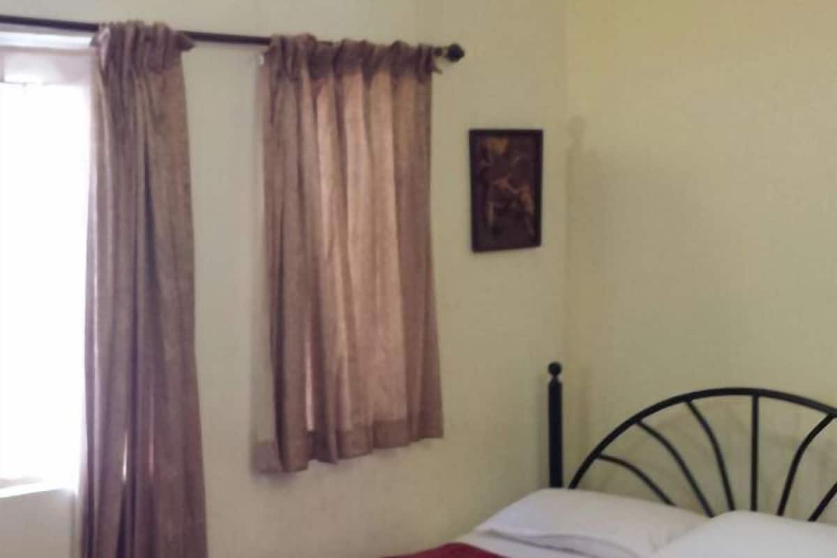 A single bed with a patterned bedspread, a ceiling fan, a window with curtains, and a wall-mounted picture.