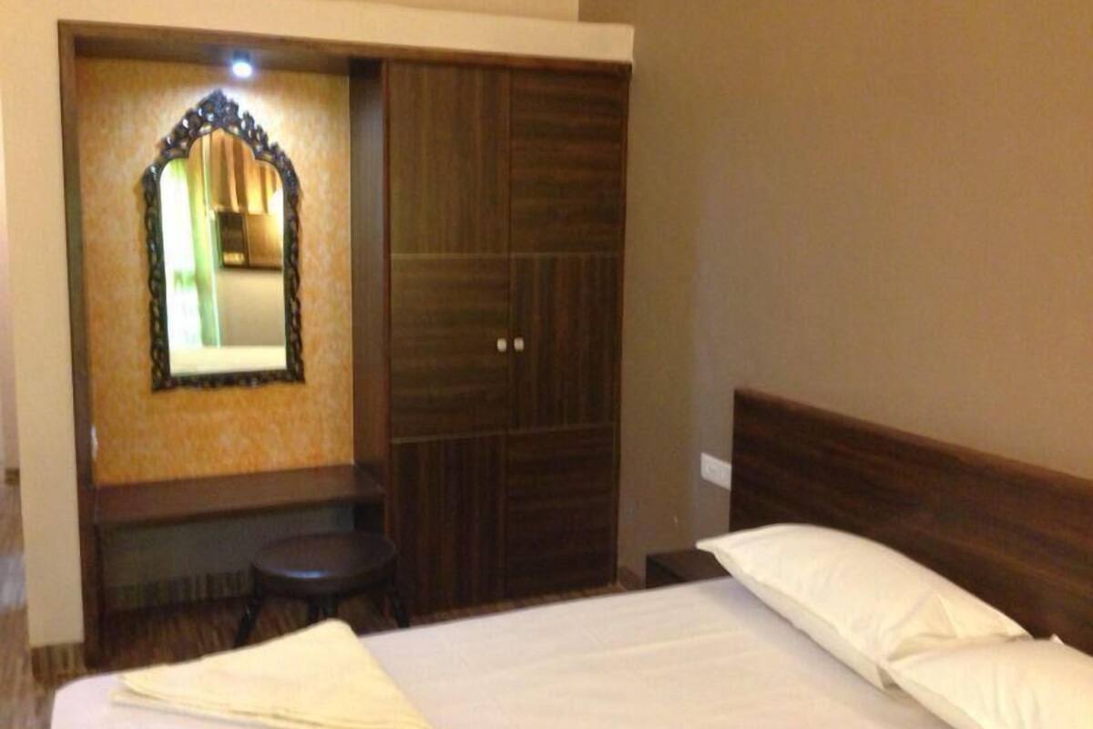 A bedroom with a bed, a wooden wardrobe, a small stool, and a mirror on the wall.
