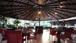 An outdoor cafe with red chairs and tables, a covered area with a metal roof, and umbrellas providing shade. - So My Resort (Calangute)