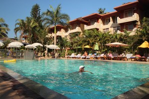 A swimming pool with lounge chairs and umbrellas, surrounded by palm trees and a hotel building. - So My Resort (Calangute)
