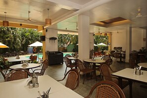 A restaurant with multiple dining tables and chairs, a pool area in the background, and ceiling fans. - Lambana Resort (Calangute)