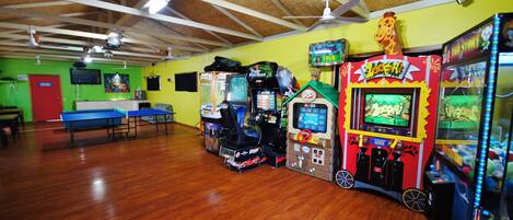 Habitación con pisos de madera, paredes verdes y ventiladores de techo. Hay máquinas de arcade y una mesa de ping pong.