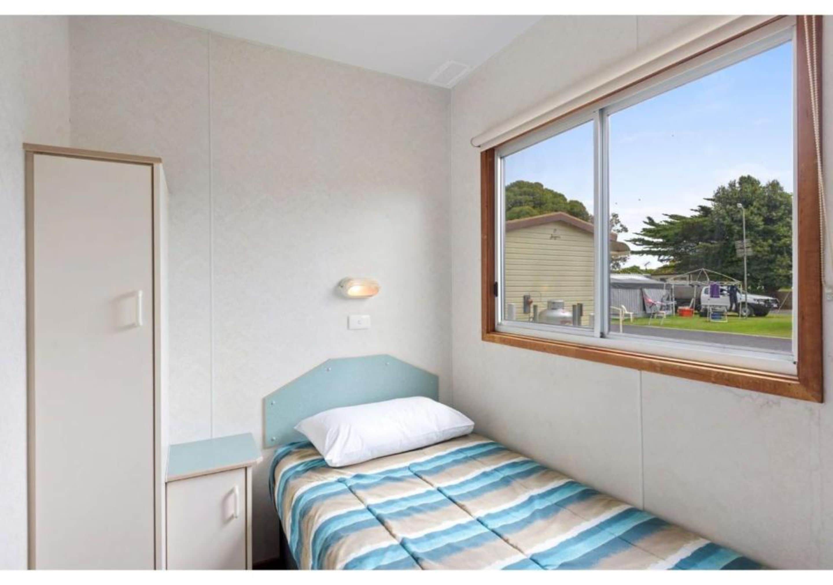 A small, single-bed room with a window, a bedside lamp, and a door.