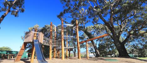 A playground with a slide, climbing net, and swings, surrounded by trees.