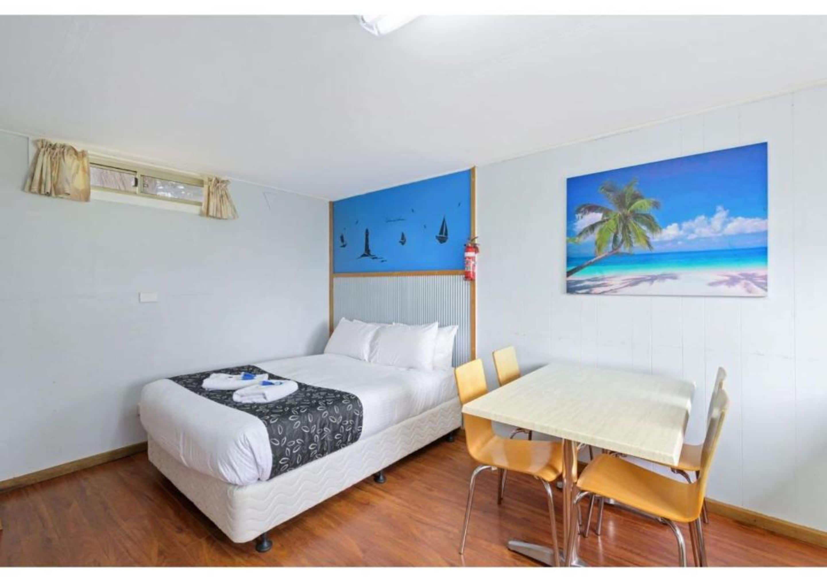 A hotel room with a bed, a dining table with chairs, a painting of a beach scene, and a window with curtains.