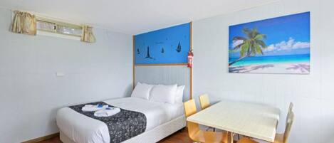 A hotel room with a bed, a dining table with chairs, a painting of a beach scene, and a window with curtains.