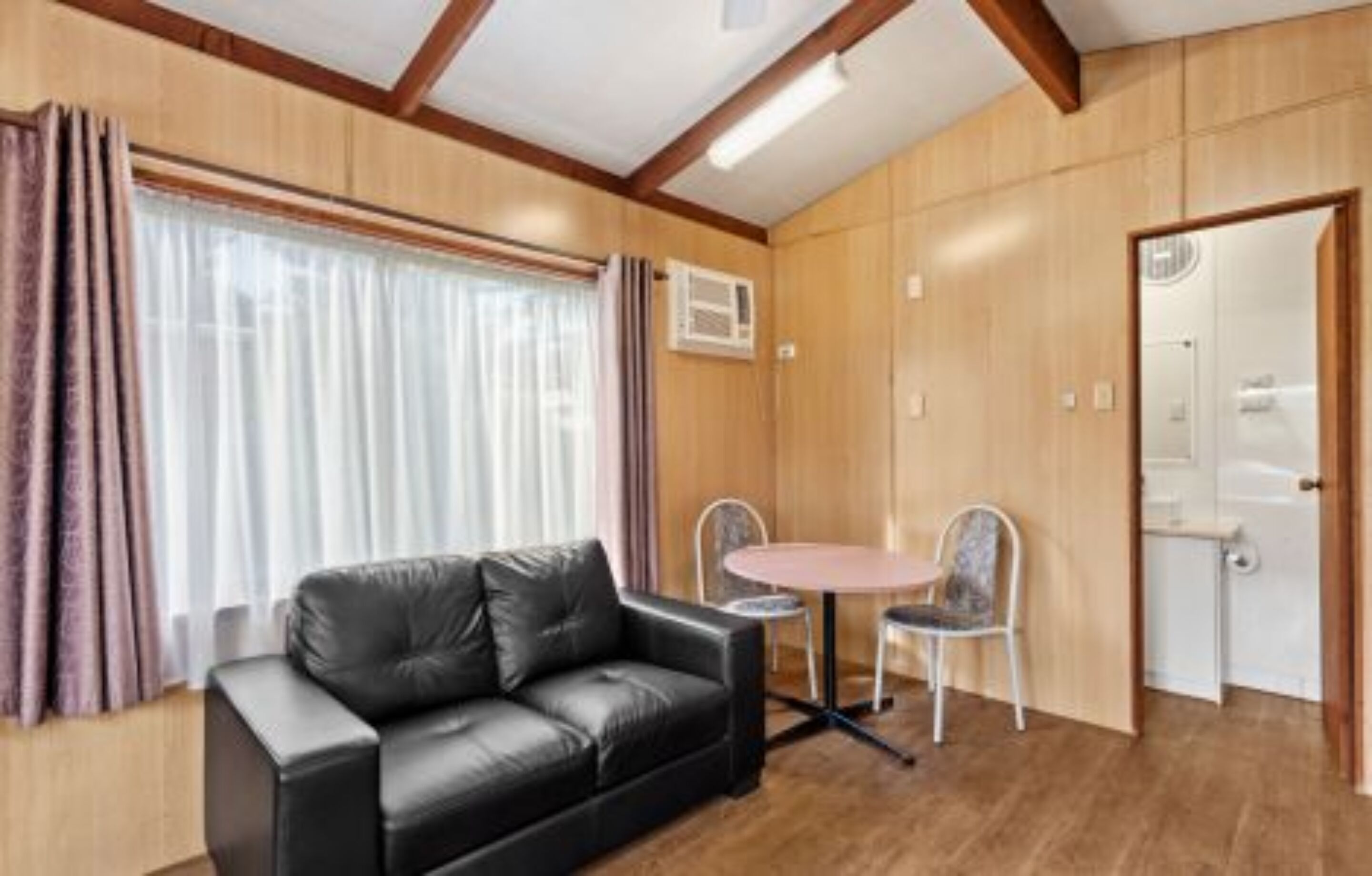 A room with a black leather sofa, a small round table with two chairs, wooden walls, and a door.