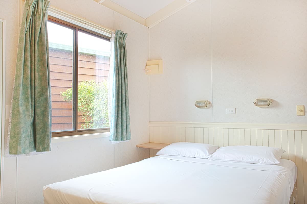 A single bed with white linens, a window with green curtains, and a wall-mounted light fixture.