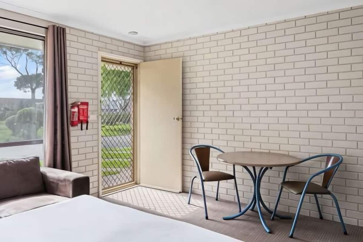 A hotel room with a bed, a table with two chairs, a window with curtains, a door, and a brick wall.