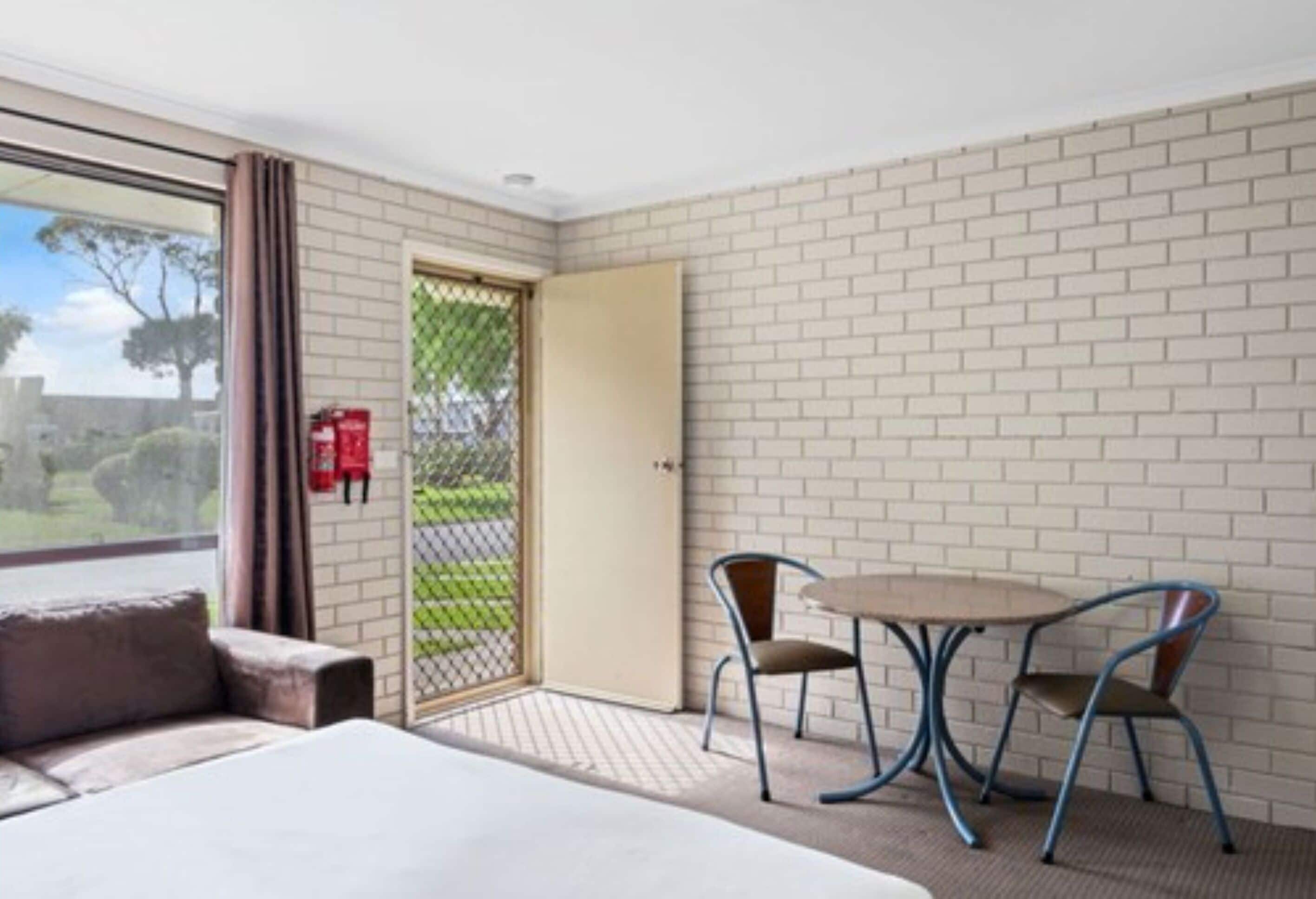 A hotel room with a bed, a small table with two chairs, a window with curtains, and a brick wall.