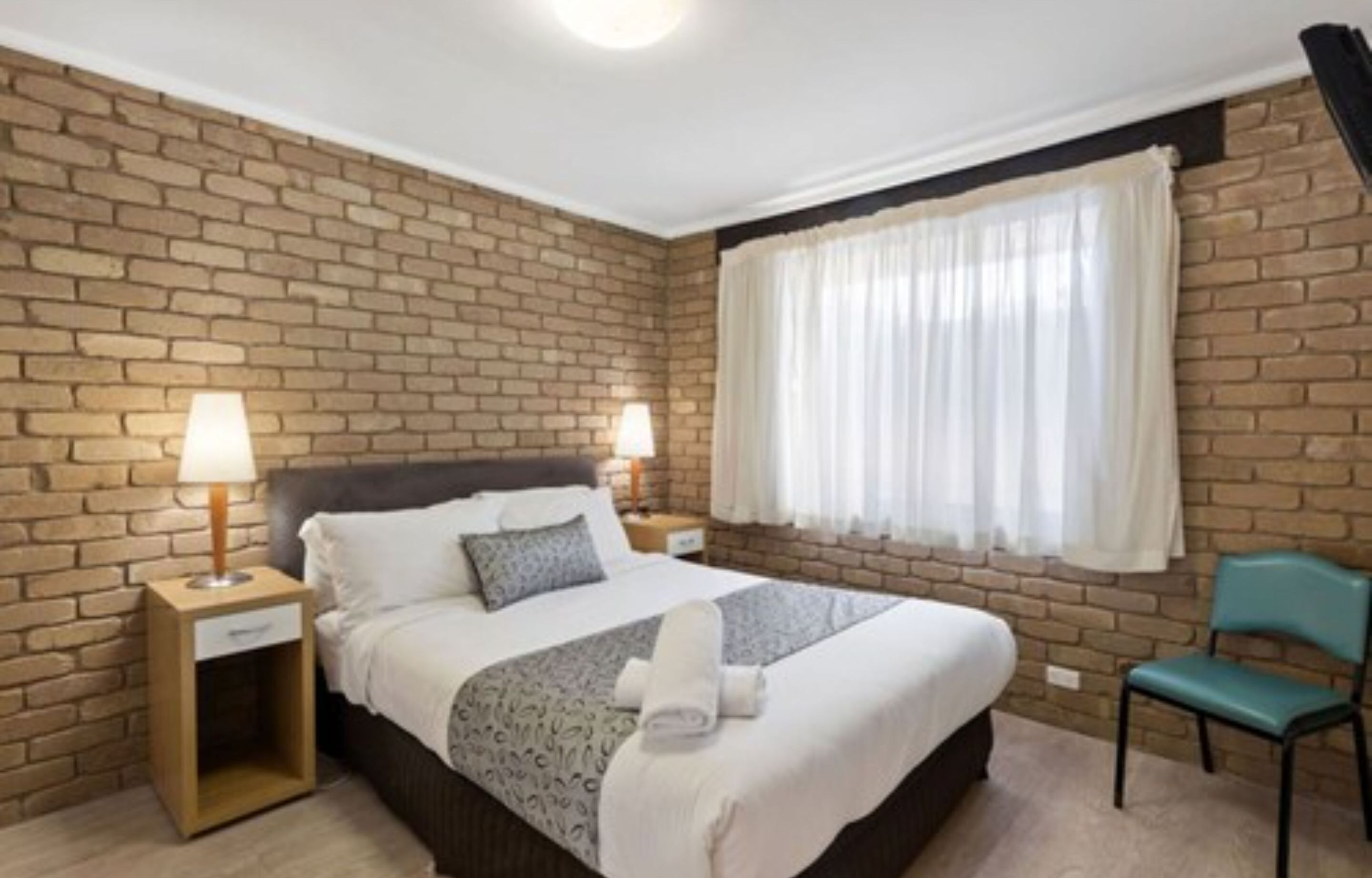 Een hotelkamer met een bakstenen muur, een bed met wit bedlinnen, twee bedlampjes, een raam met witte gordijnen en een teal-kleurige fauteuil.