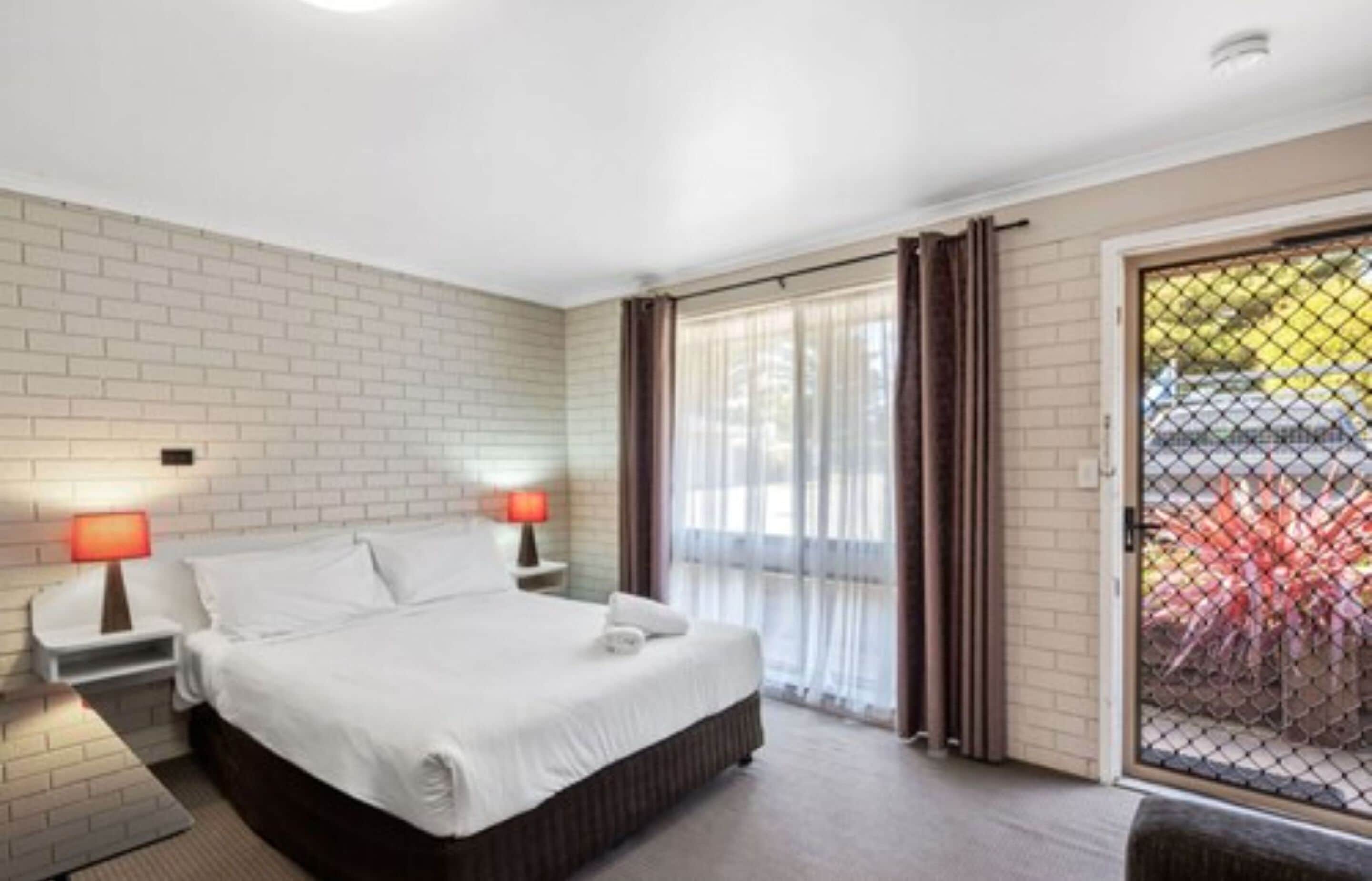 A hotel room with a bed, two bedside lamps, a window with curtains, and a view of the outside.