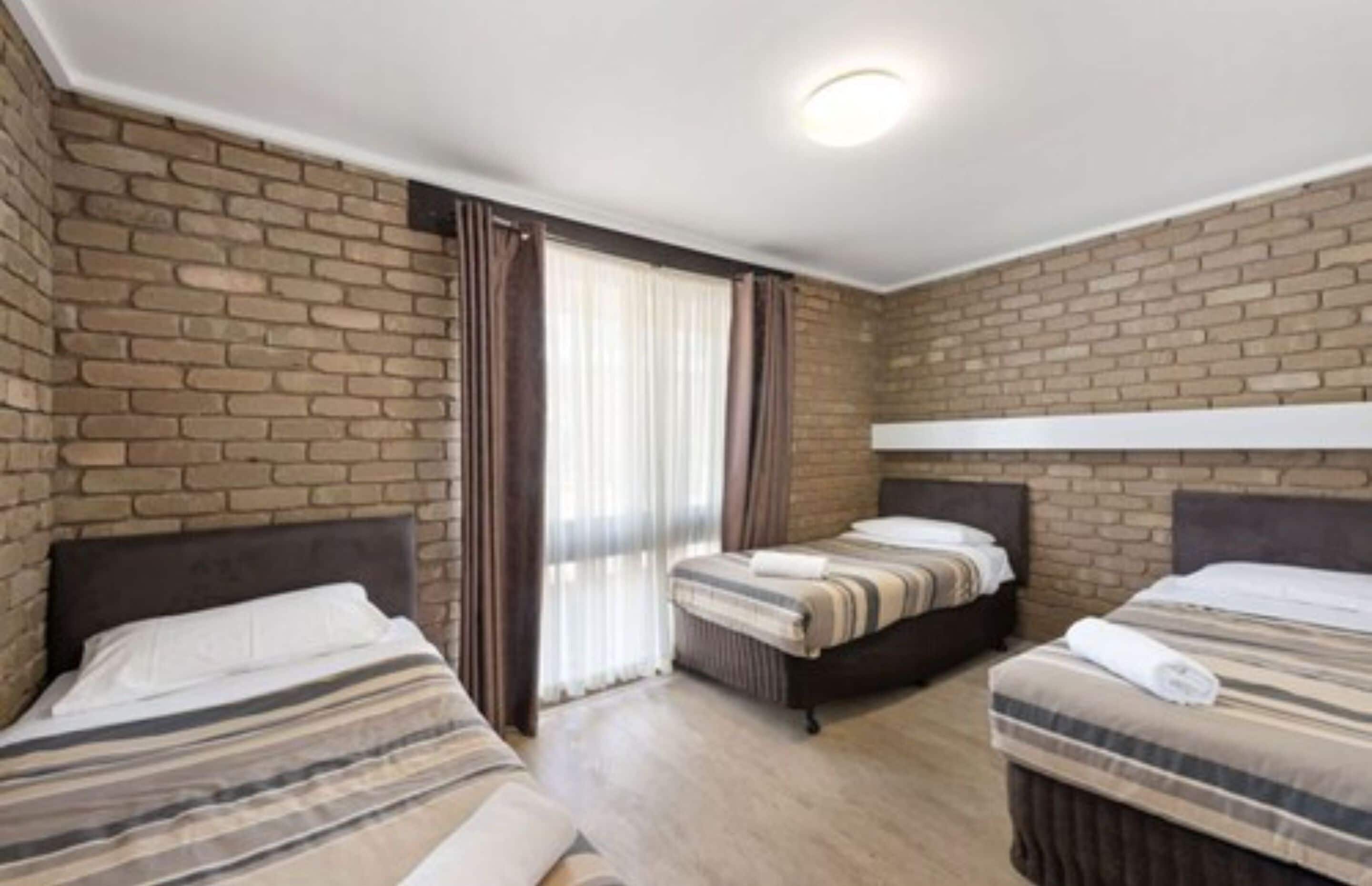 A hotel room with two beds, a brick wall, and a window with curtains.
