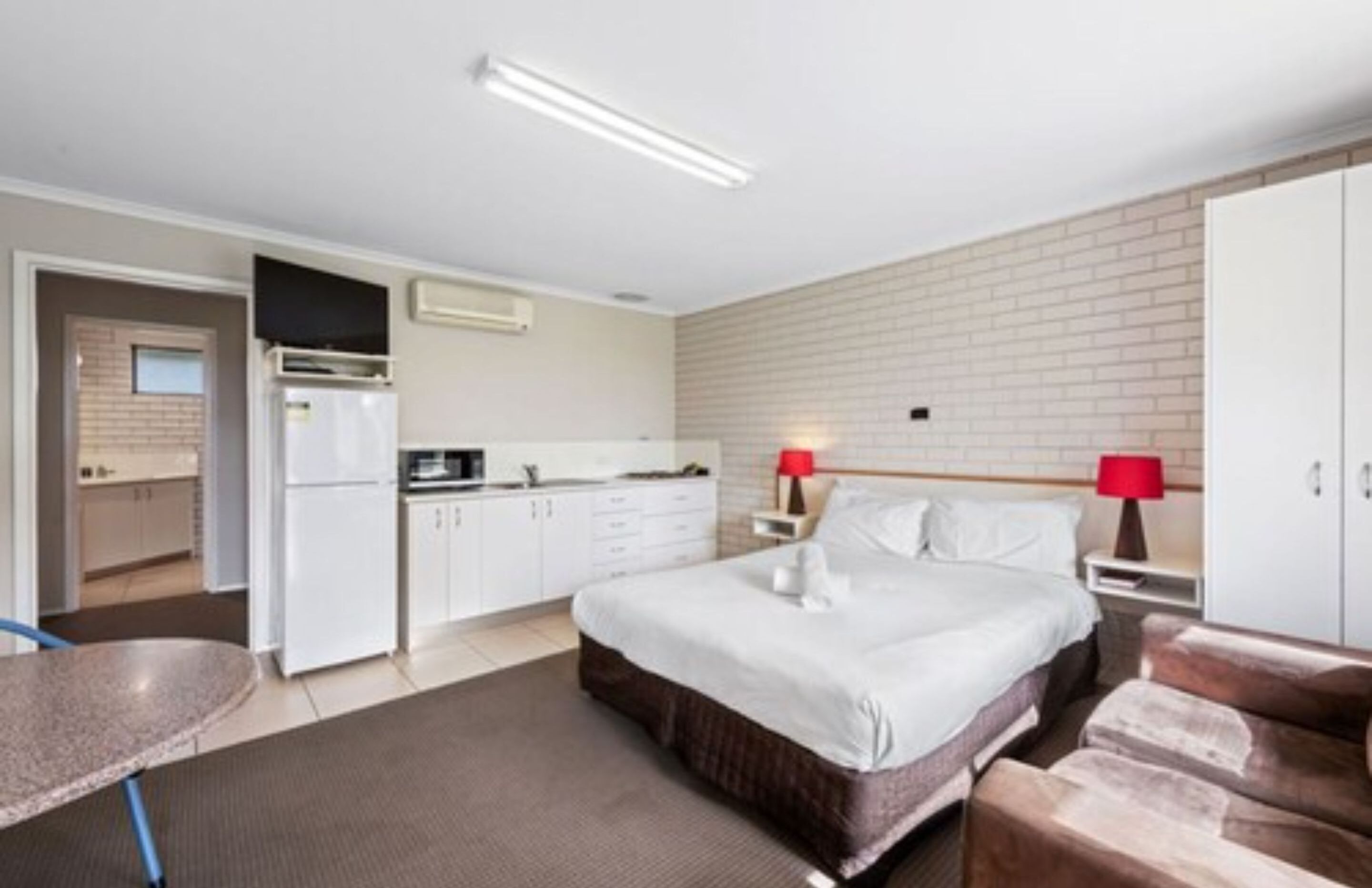 A hotel room with a bed, bedside tables, a red lamp, a kitchenette, and a bathroom visible through an open door.