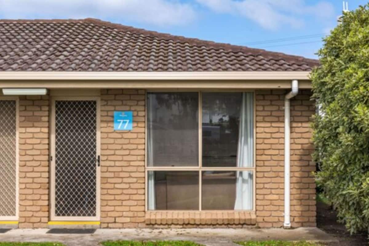 A single-story building with a brown tiled roof and beige brick walls. There is a large window on the front and a door with a metal mesh security screen. A blue sign with the number 77 is visible on the wall.