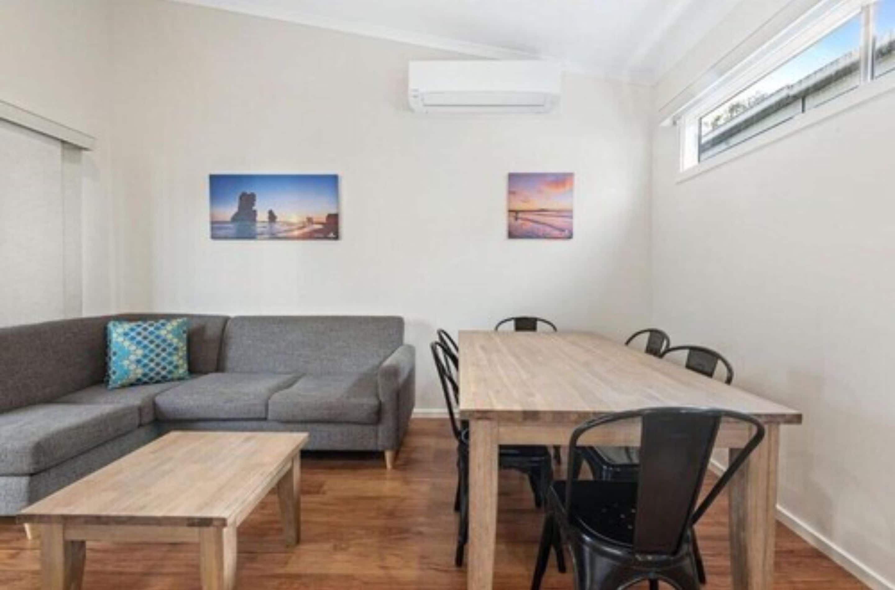 A modern living room with a grey sofa, a wooden dining table with chairs, and two framed pictures on the wall.