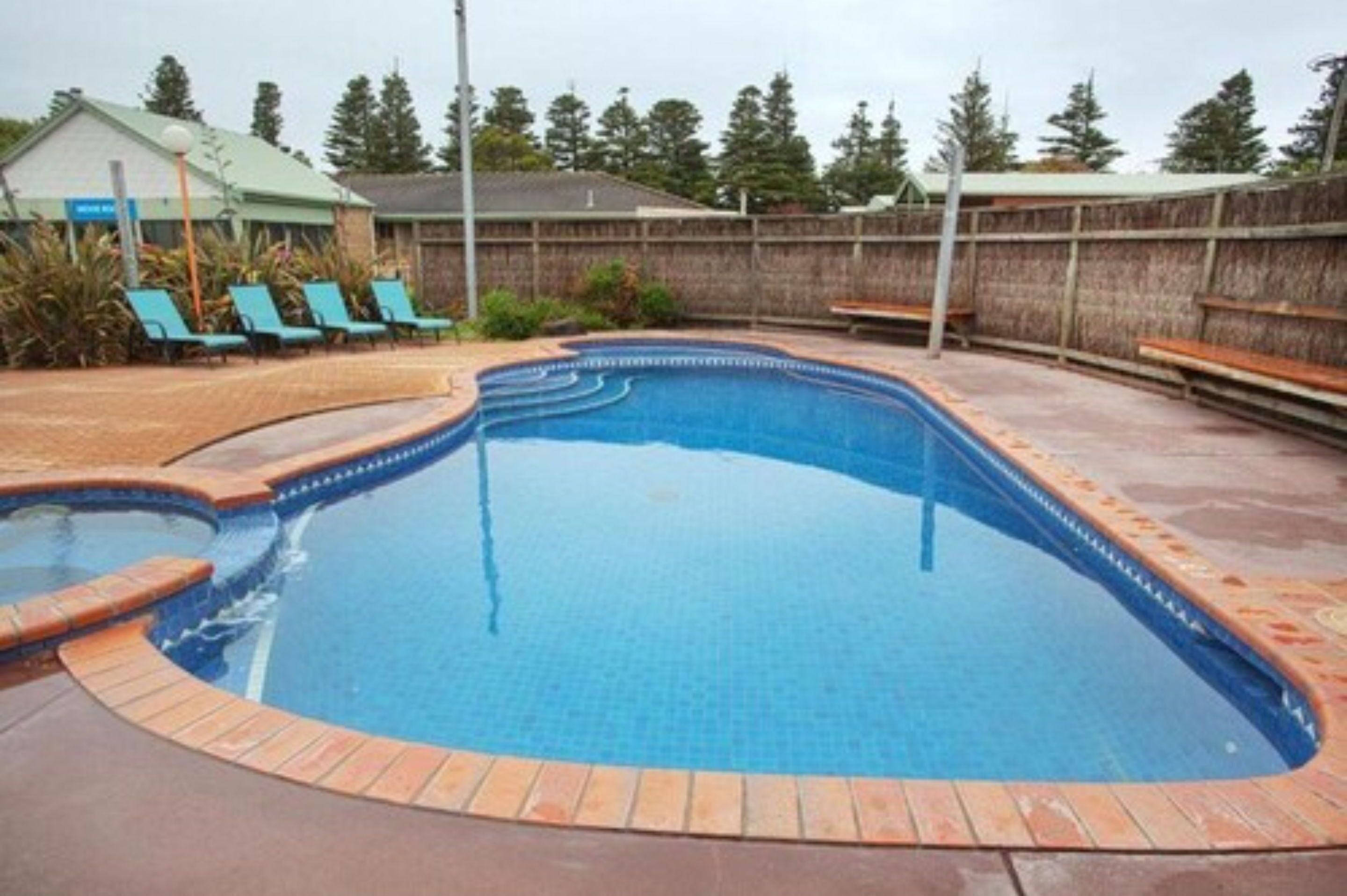 A swimming pool with a tiled edge, surrounded by a brick deck and a wooden fence.