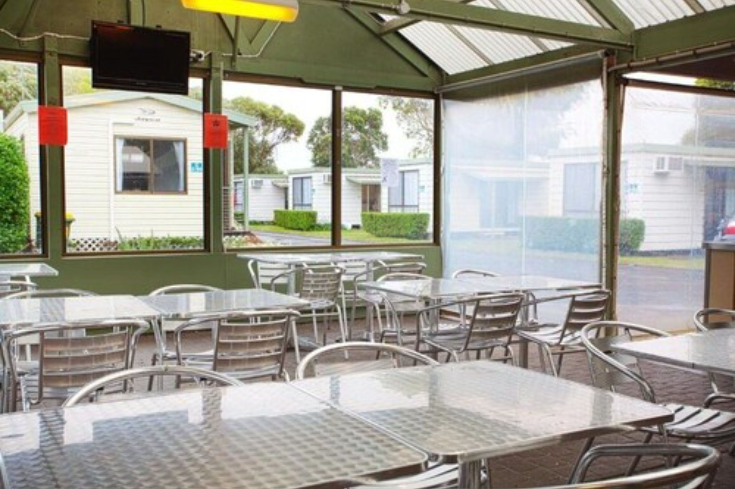 A covered outdoor dining area with metal tables and chairs, large windows, and a television screen.
