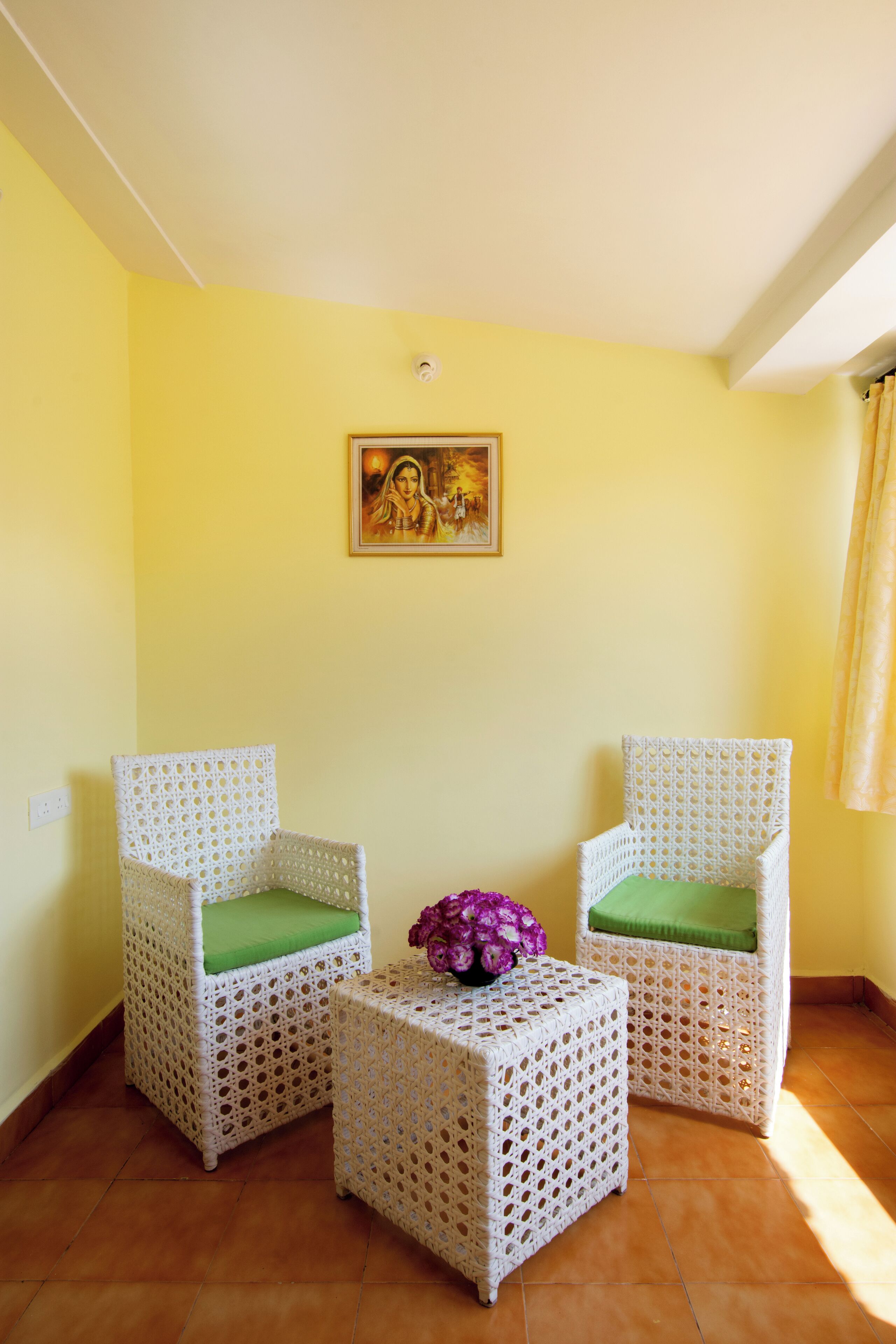 A room with two wicker chairs with green cushions, a small table with a purple flower, and a wall-mounted picture.