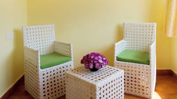 Quarto com duas cadeiras de vime com almofadas verdes, uma pequena mesa com uma flor roxa e um quadro pendurado na parede.