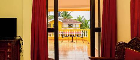 A room with a television on a wooden cabinet, a red sofa, and a view of a yellow building with a tiled patio and a white fence.