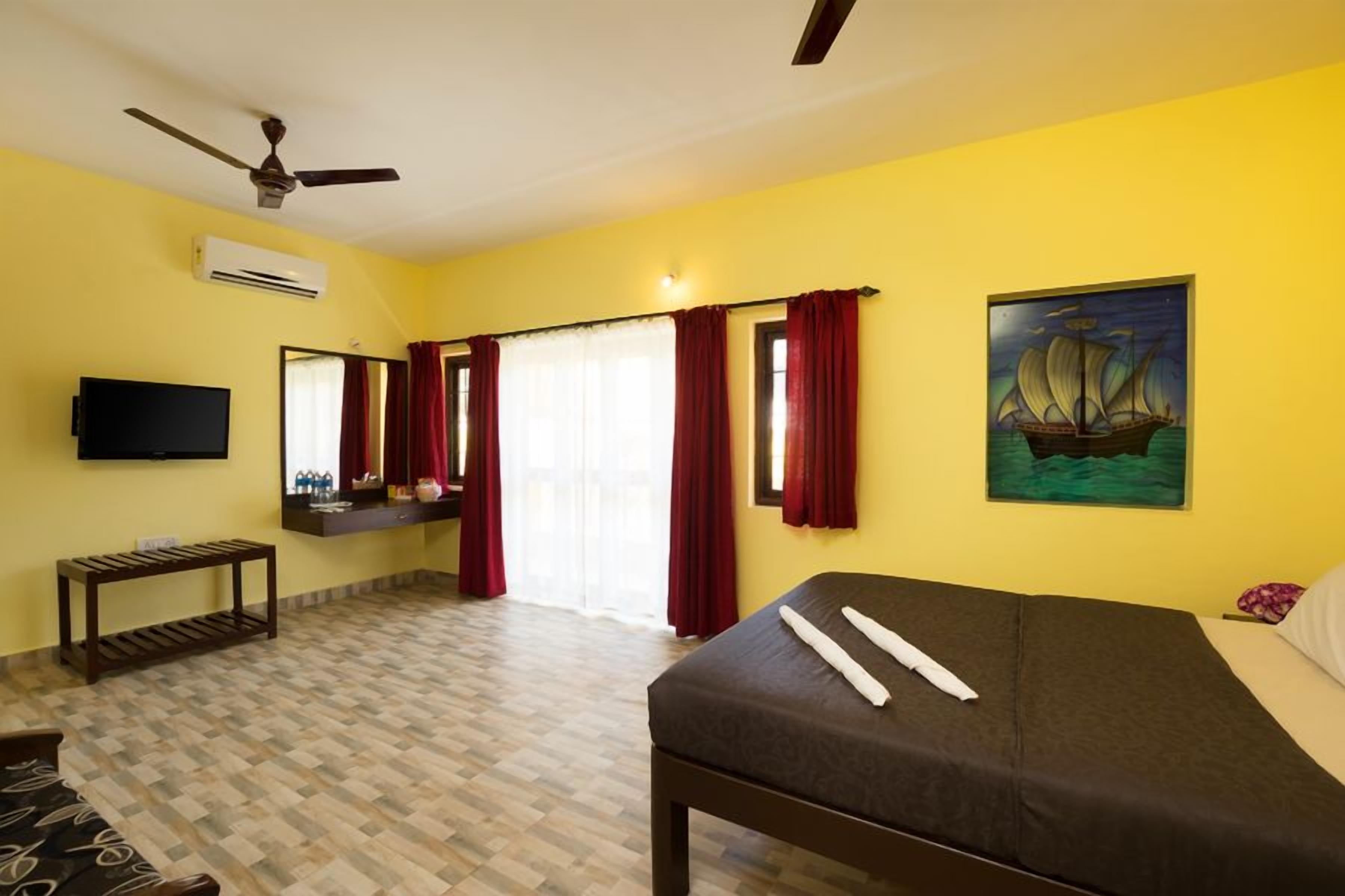 Habitación de hotel con cama, televisión, ventilador de techo, ventana con cortinas rojas y un cuadro en la pared.