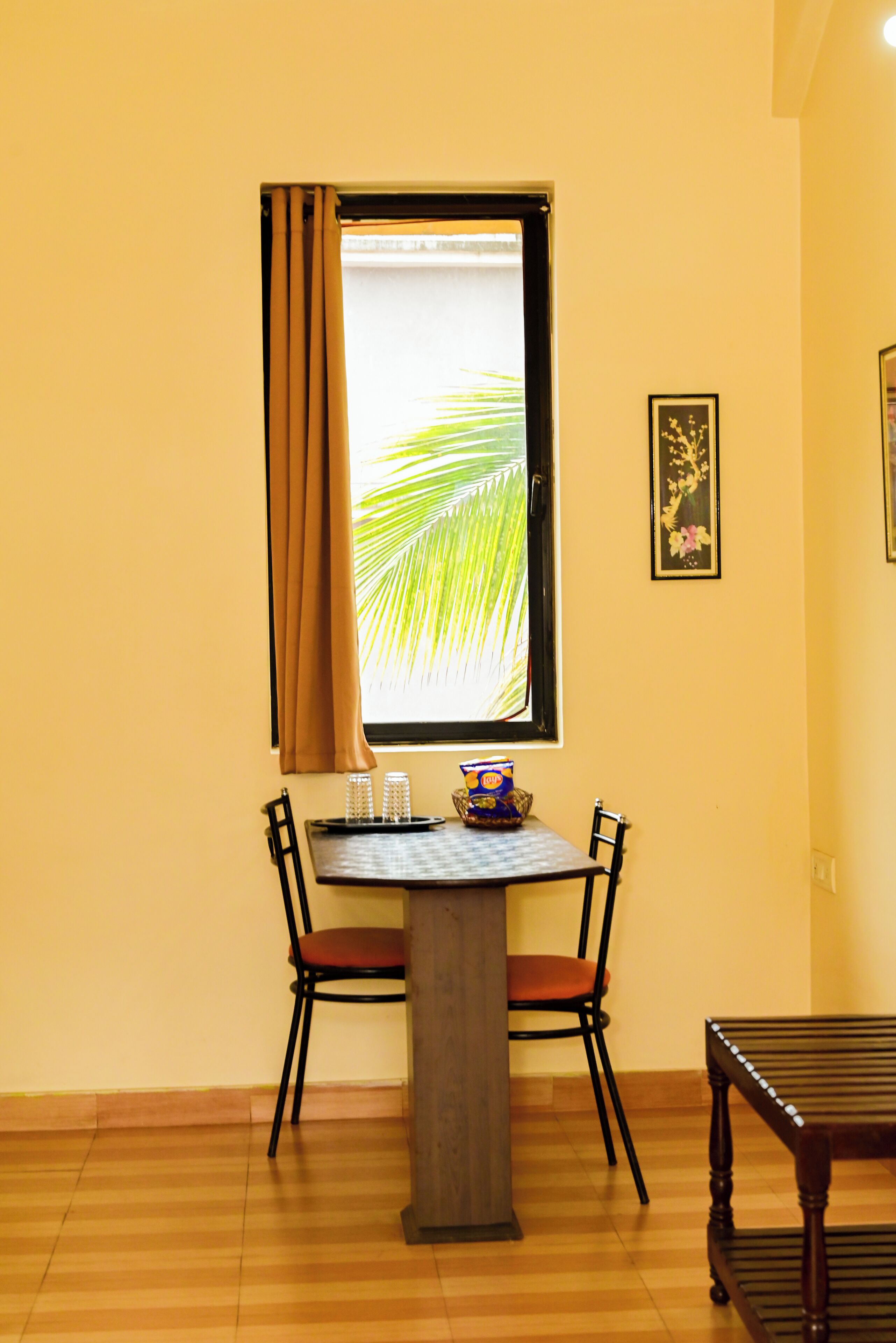 A room with a dining table, two chairs, a bench, a window with curtains, and a framed picture on the wall.