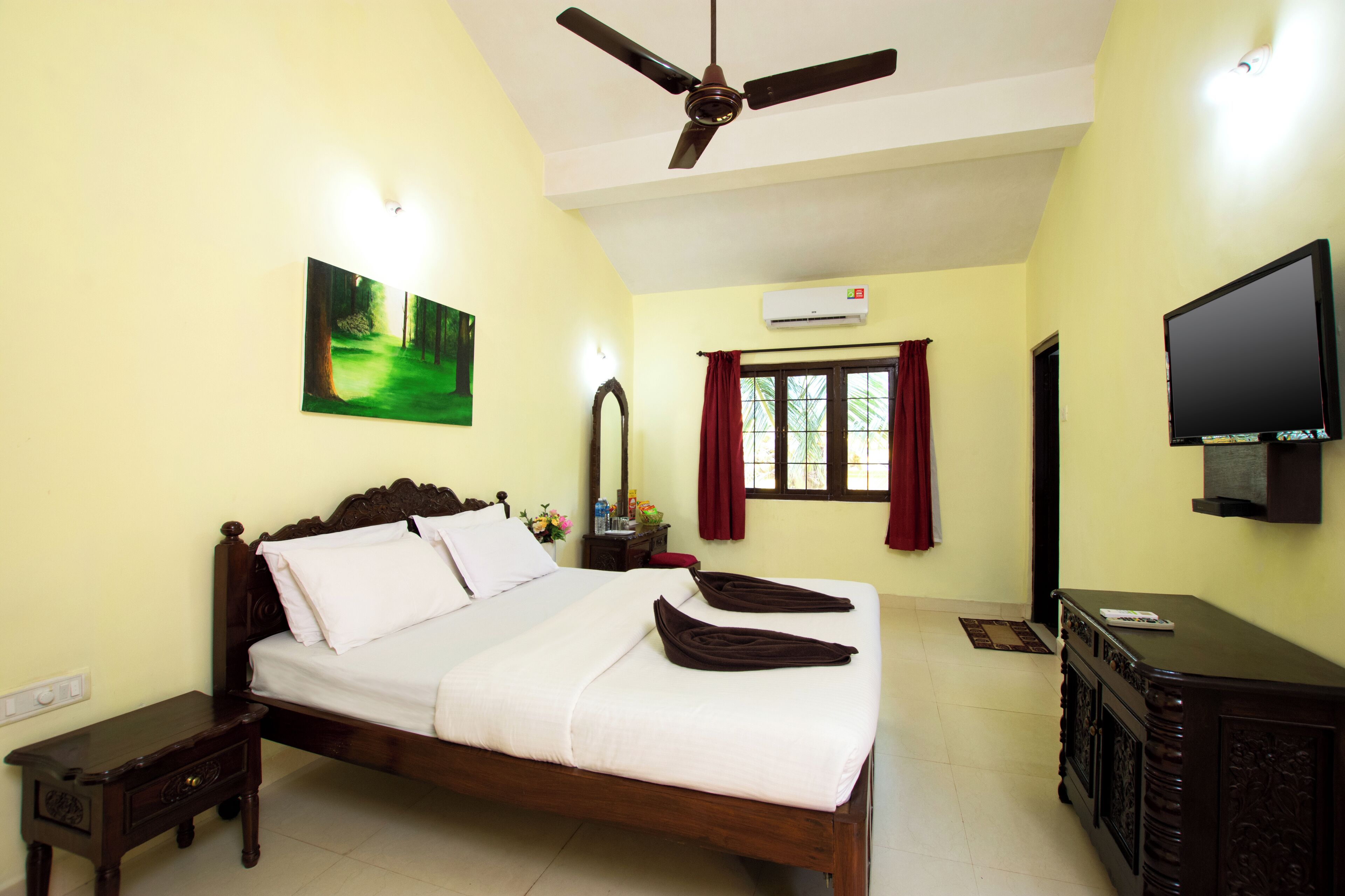 A hotel room with a bed, a ceiling fan, a flat-screen TV, a painting, and an air conditioning unit.