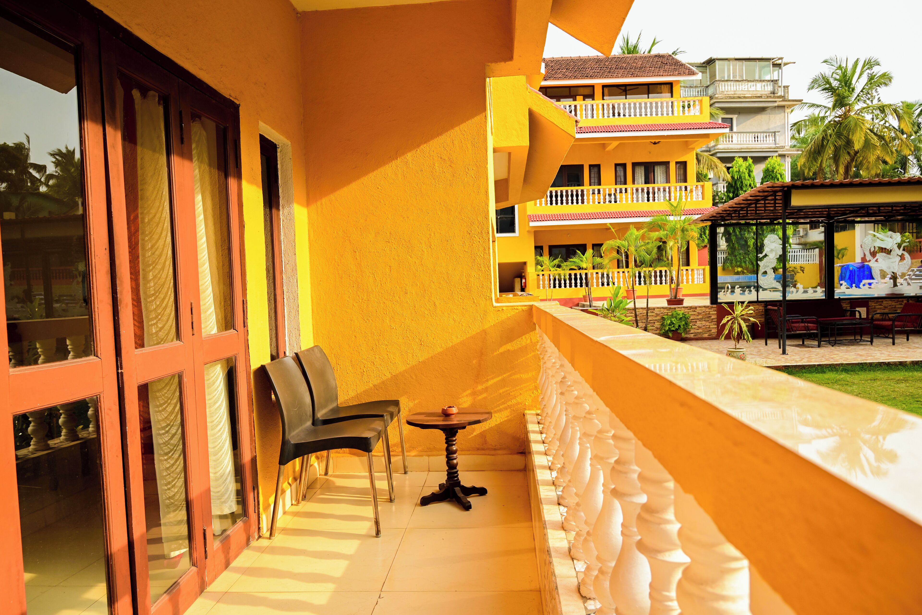 A balcony with a table and chairs, a view of a building with balconies, and a small garden area.