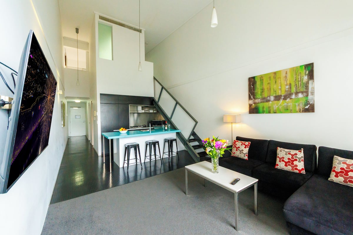 A modern living room with a black sofa, a white coffee table, and a kitchen area with a bar and stools.