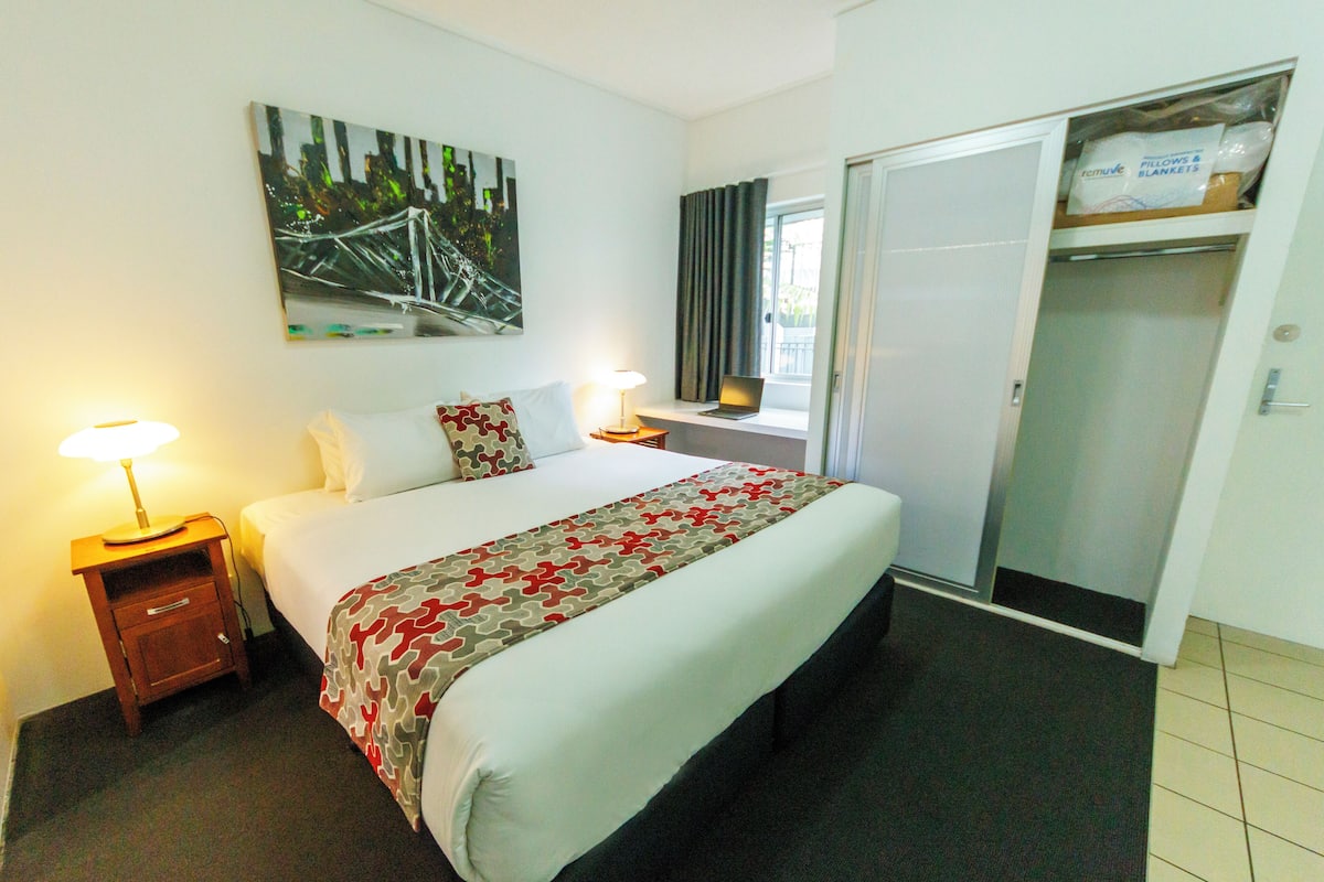 A hotel room with a bed, bedside lamps, a window with curtains, a laptop on the windowsill, and a wardrobe with a mirror.