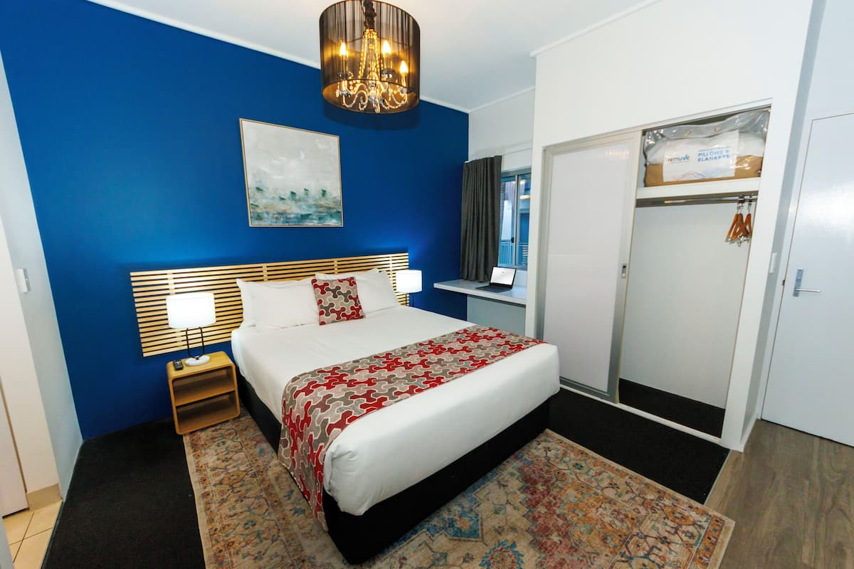 A hotel room with a large bed, a nightstand, a chandelier, a painting, a wardrobe, and a window with curtains.