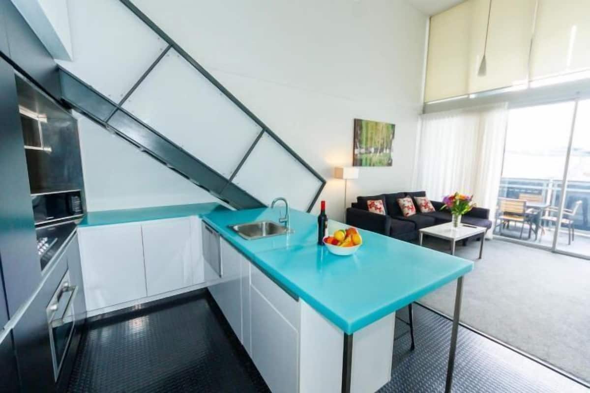 A modern kitchen with a blue countertop, white cabinets, and a black floor. There is a dining area with a black sofa and a table with a vase of flowers.