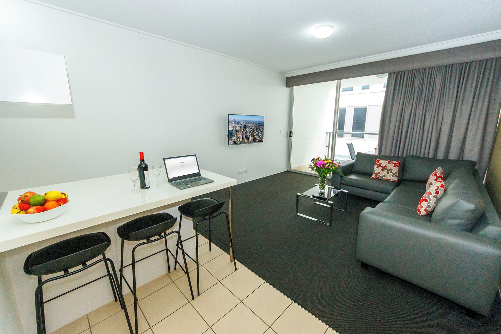 A modern living room with a grey sofa, a dining area with a white table and black chairs, a laptop, and a fruit bowl.