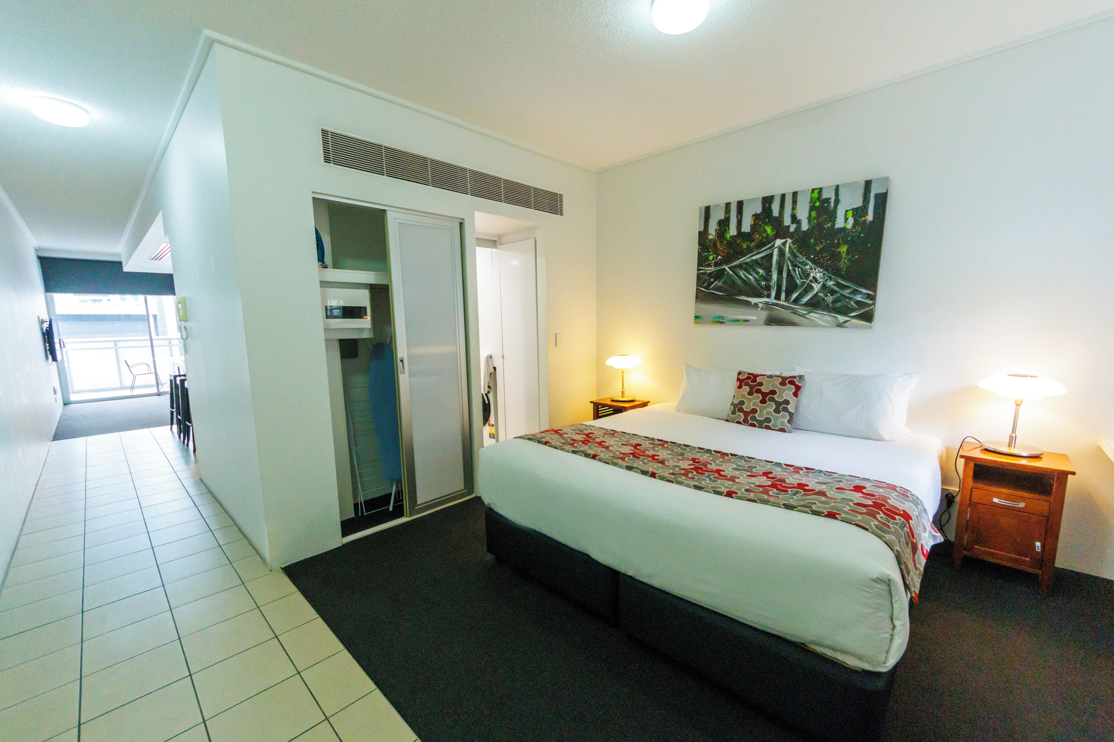 A hotel room with a large bed, a wardrobe, a bedside table with a lamp, and a painting on the wall.